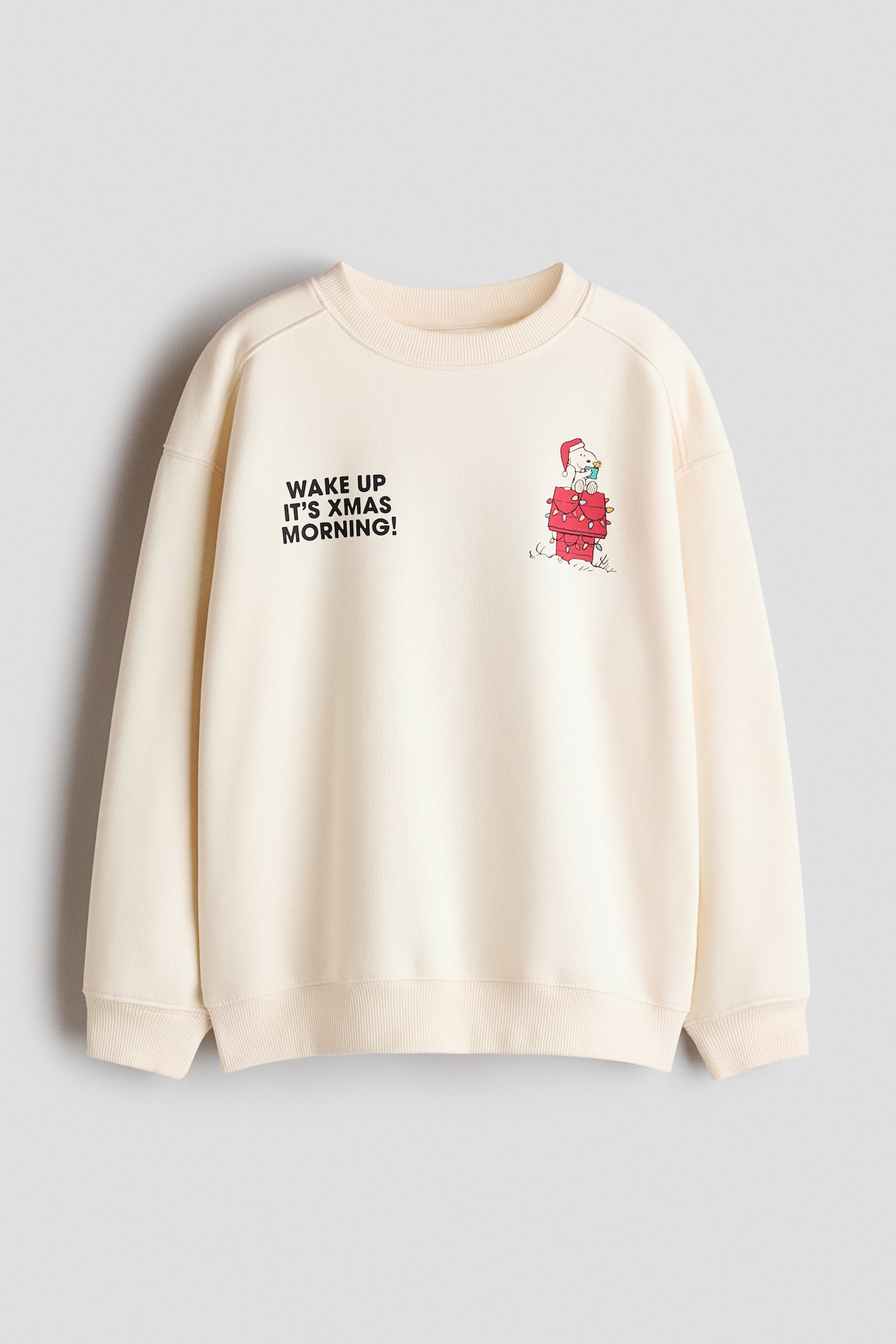 A relaxed-fit, off-white sweatshirt is shown from the front, featuring a round rib-trimmed neckline, dropped shoulders, and ribbed cuffs and hem. The front showcases the printed black text "WAKE UP IT'S XMAS MORNING!" on the left chest and a cartoon dog wearing a Santa hat atop a red chimney on the right chest.