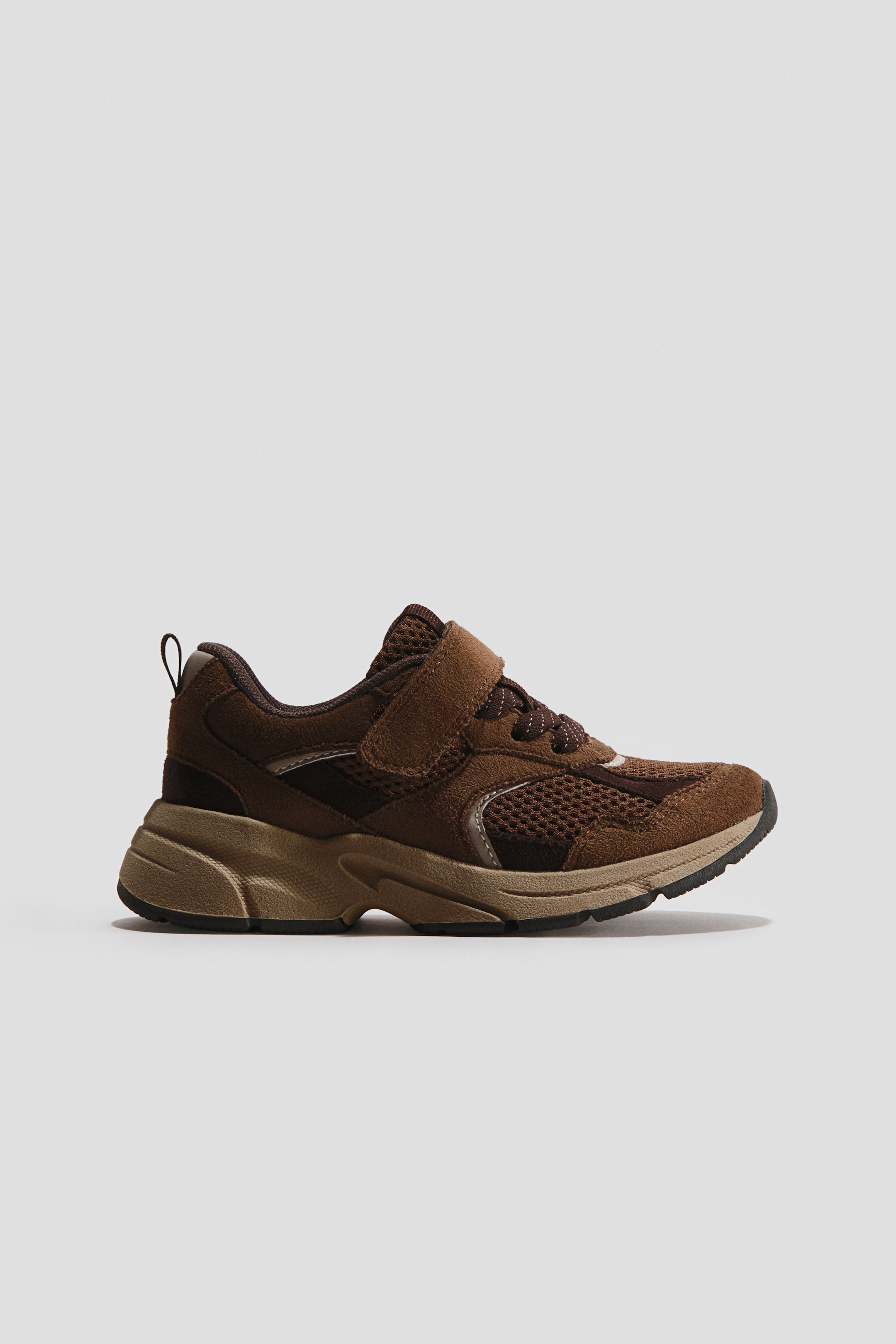 View larger image: Sneakers. A brown sneaker is displayed in a side profile, showcasing a blend of brown suede-like panels and mesh inserts. It features a Velcro strap securing over the laces and a chunky, multi-toned sole in shades of brown and tan.