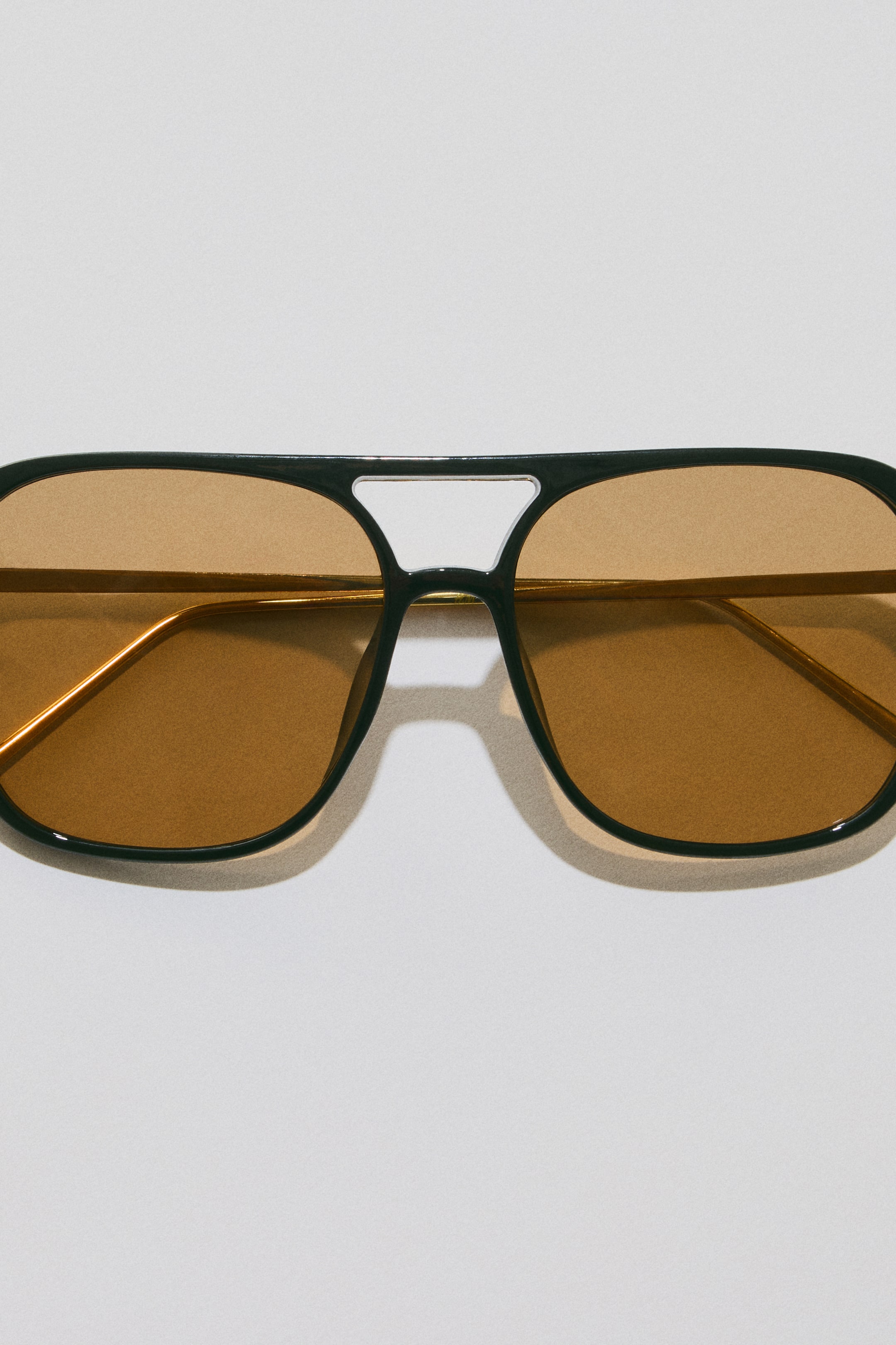Agrandir l'image: Les lunettes de soleil présentent une monture de style aviateur noire avec des verres teintés marron. Un double pont forme le sommet de la monture, et des branches fines de couleur or sont visibles à travers les verres, les lunettes étant orientées vers l'avant et légèrement inclinées vers le bas.