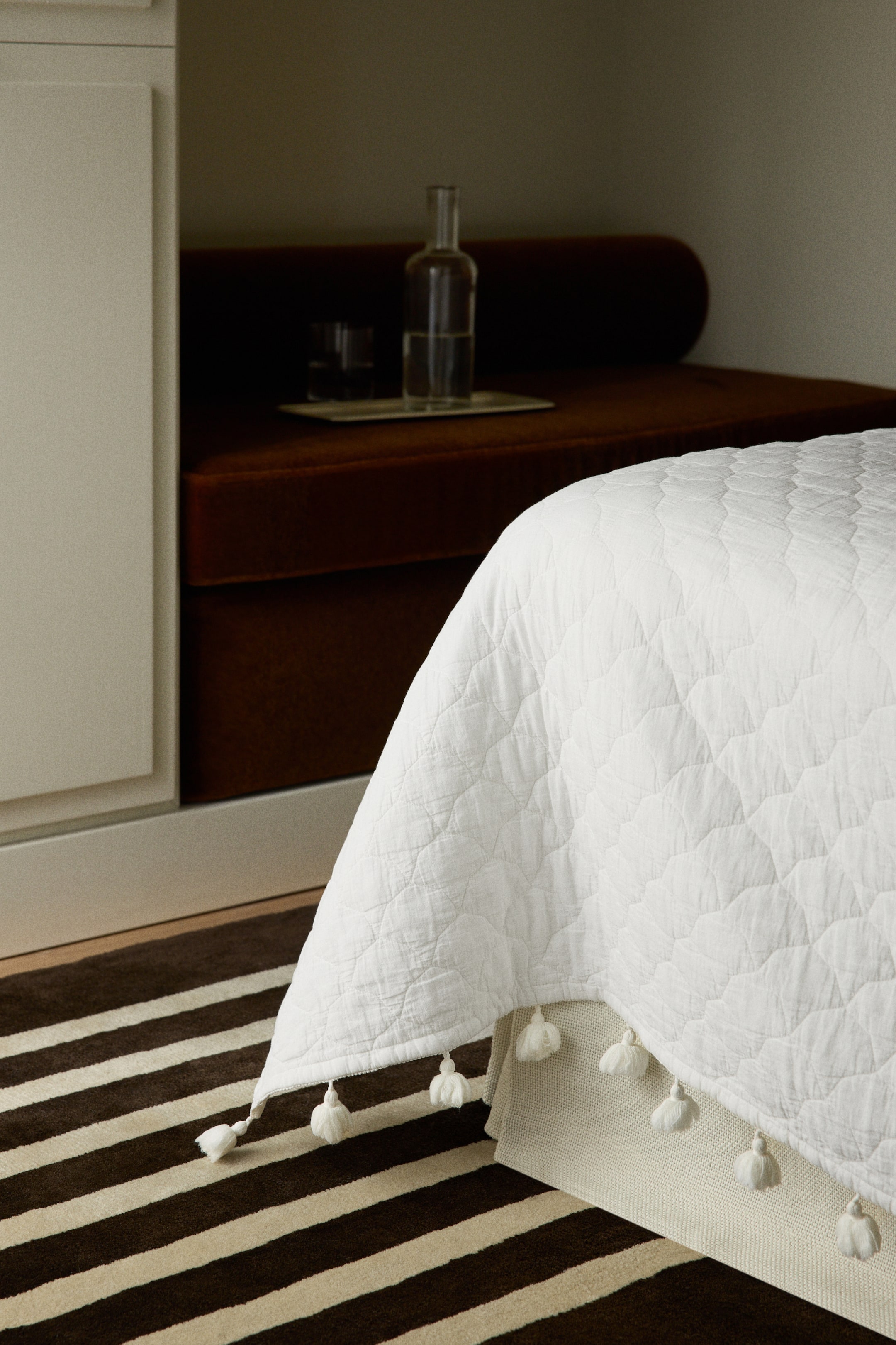 Ingrandisci l'immagine: Un copriletto trapuntato bianco presenta un sottile motivo geometrico e sfoggia nappe bianche lungo il bordo inferiore, visto da un'angolazione laterale-frontale.