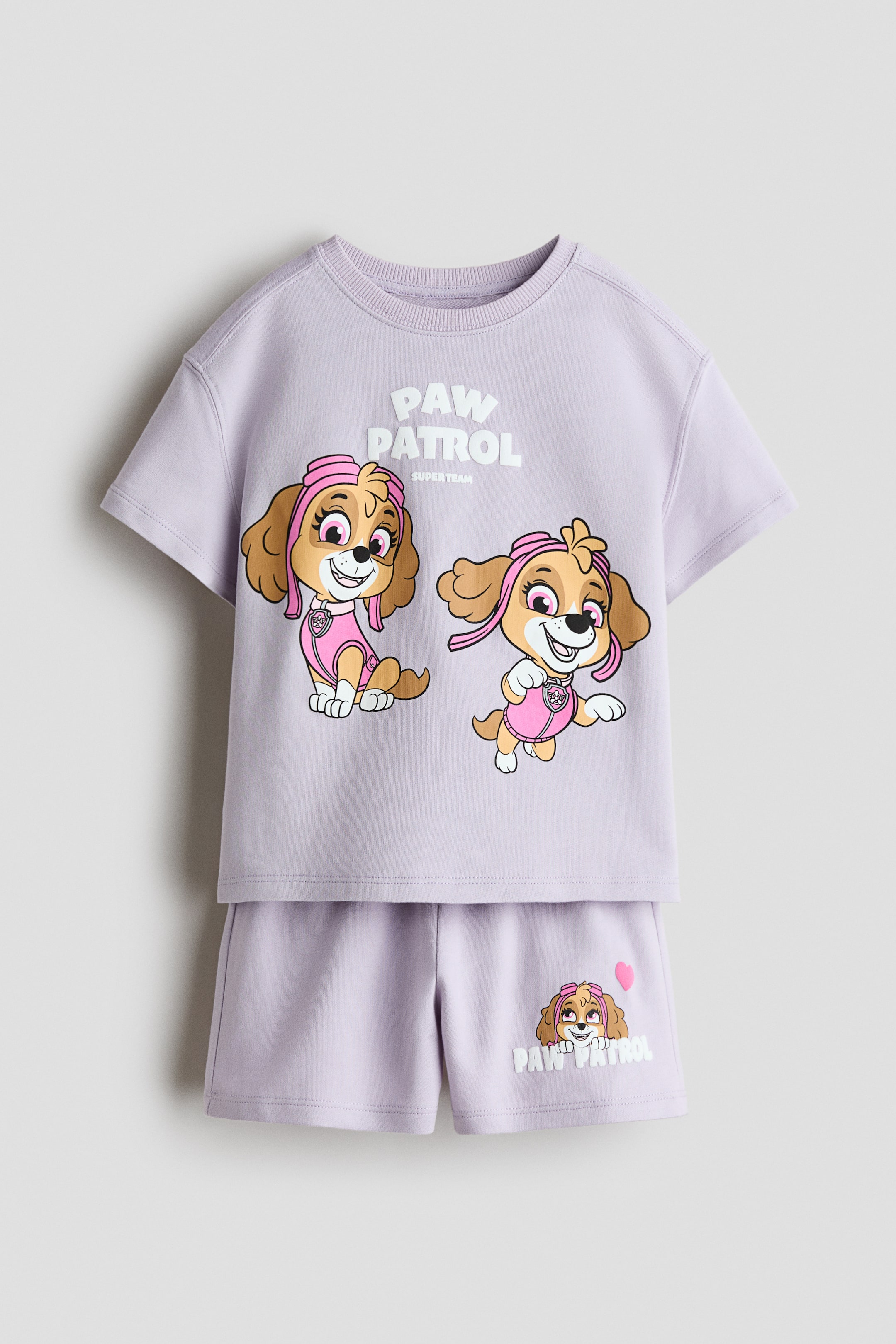 View larger image: A light purple two-piece children's set showcases a short-sleeved top and matching shorts, front-facing. The top features a ribbed crew neck and two cartoon dog characters adorned with pink accessories, with the text "PAW PATROL SUPER TEAM" displayed above them. The shorts highlight a smaller cartoon dog character's head, "PAW PATROL" text, and a pink heart on the left leg.