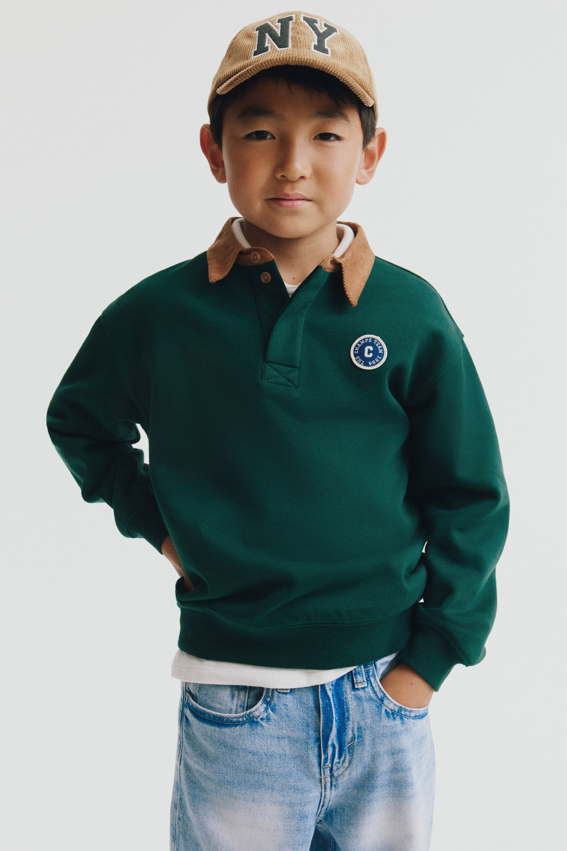 Visa större bild: En grön rugbyskjorta visas framifrån och har en kontrasterande krage i brun manchester, knappslå och en cirkelformad broderad patch på vänster bröst. Sweatshirten är stylad med sänkta axlar, ribbade muddar och ribbad fåll, lager-på-lager över en vit T-shirt.