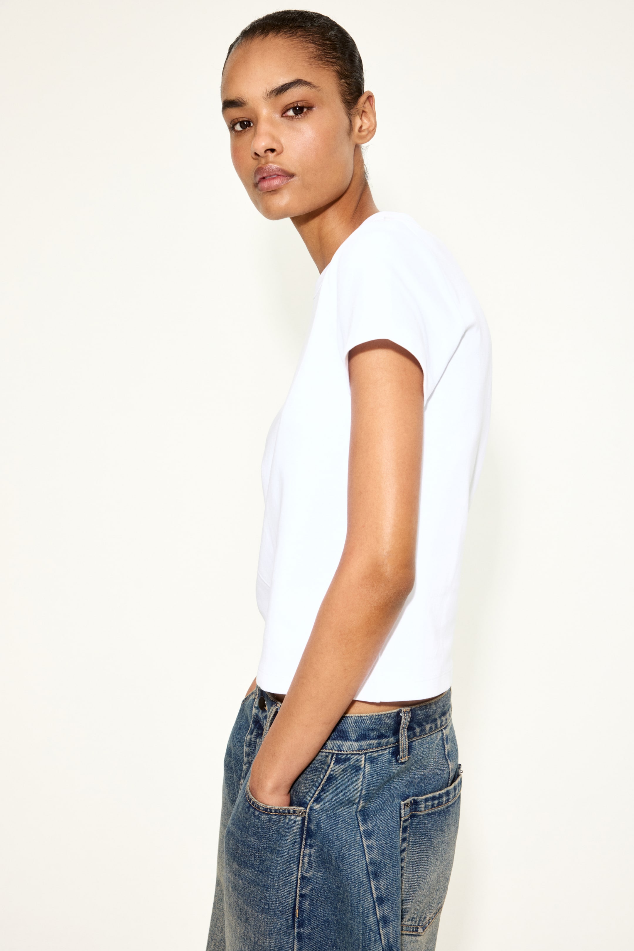 Se større billede: En hvid t-shirt med korte ærmer og en lige kant, præsenteret fra en sideprofil og slutter lige over linningen på blå jeans.