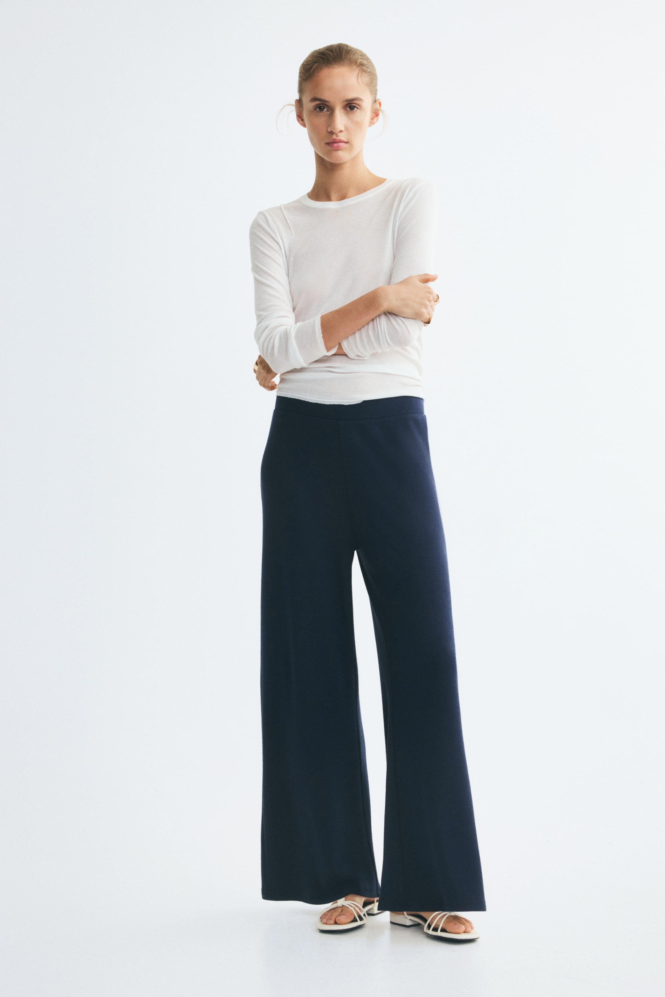 View larger image: The model is wearing loose-fit pants in a dark blue ribbed jersey, featuring a regular elasticated waist and straight legs. The pants are styled with a white long-sleeve T-shirt and white strappy sandals.