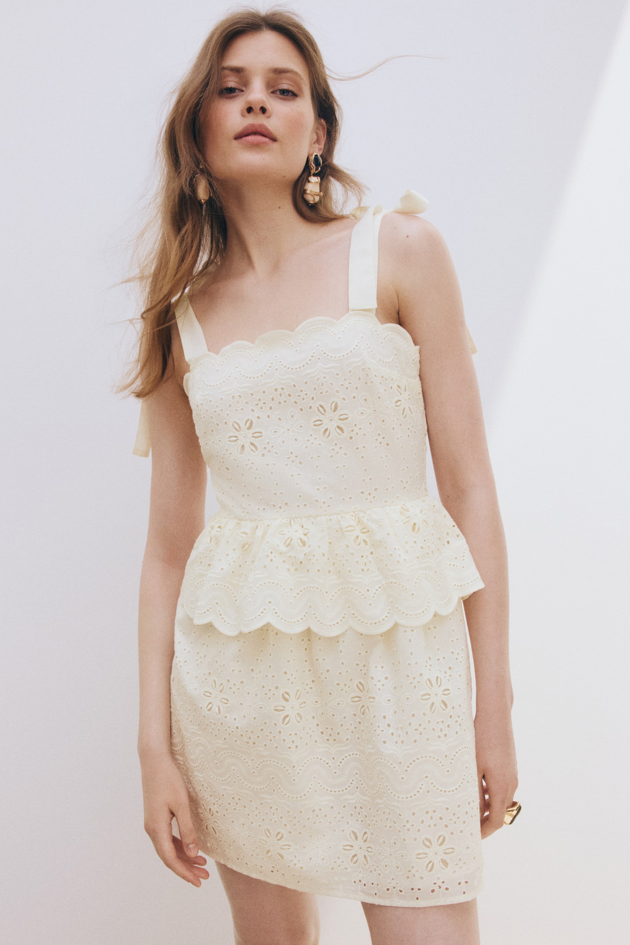 View larger image: Presenting a front-facing cream eyelet lace mini-dress, this piece features wide shoulder straps tied into bows and a scalloped neckline. The dress showcases floral embroidery throughout, a peplum waist with scalloped edges, and a matching short skirt.