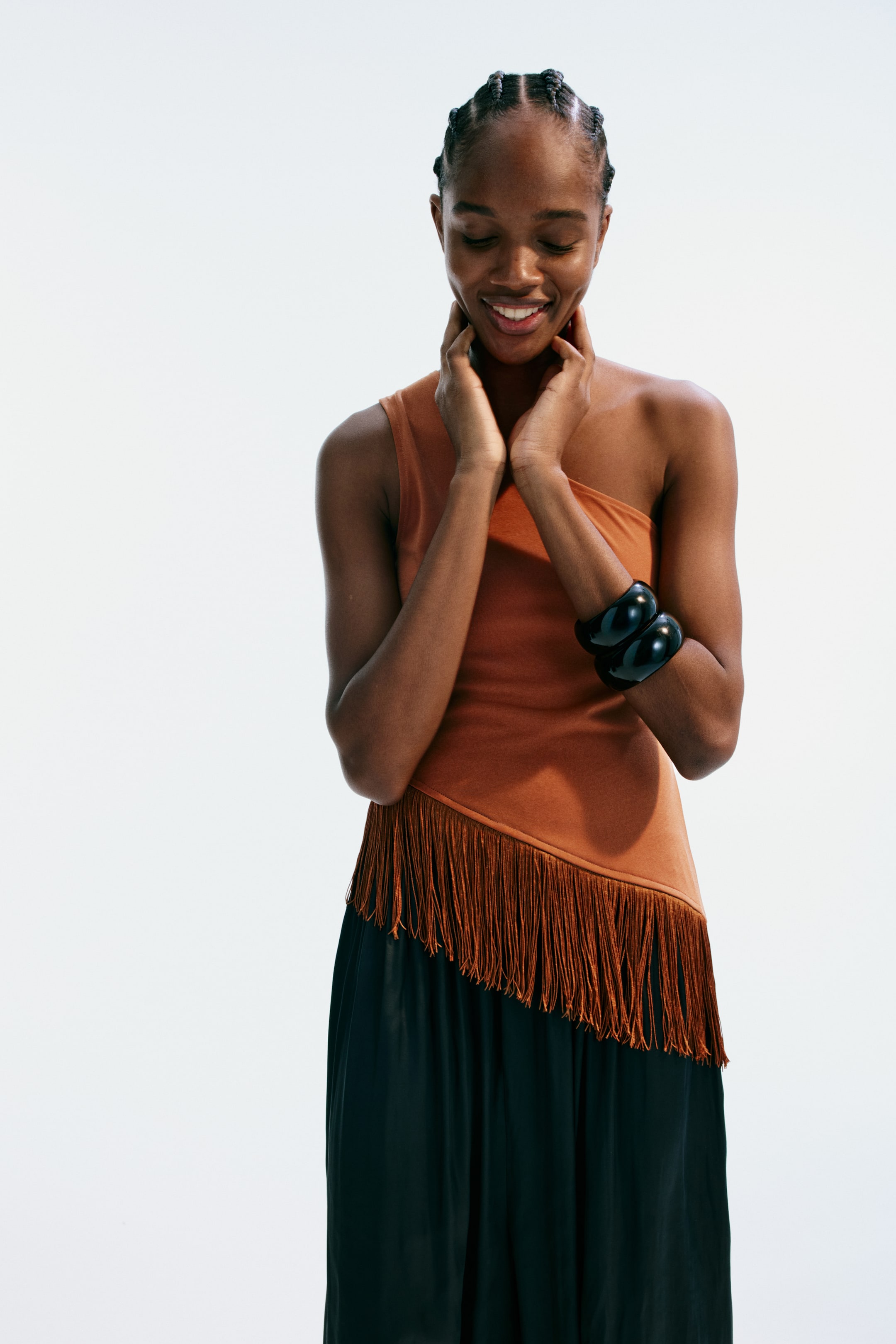 View larger image: A terracotta one-shoulder top showcases an asymmetrical hemline adorned with fringe detailing. It's paired with a dark wide-leg skirt, complemented by two black bangles on the right wrist.