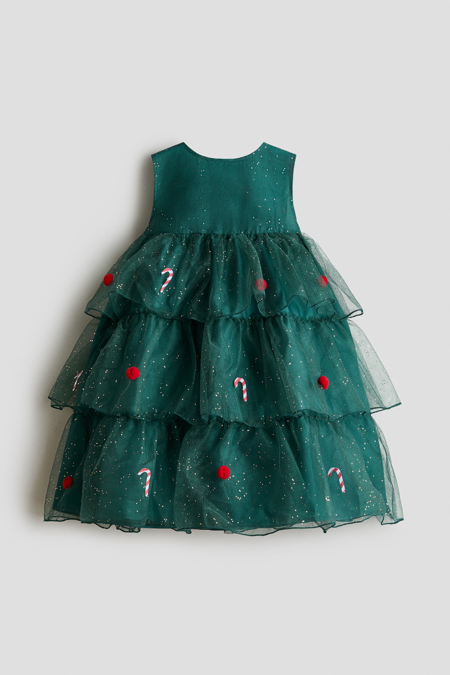 A green sleeveless dress is shown from the front, featuring a three-tiered ruffled tulle skirt embellished with gold glitter, small red pom-poms and red-and-white striped candy cane motifs. The dress has a round neckline and a voluminous, layered silhouette created by the sheer tulle overlays.