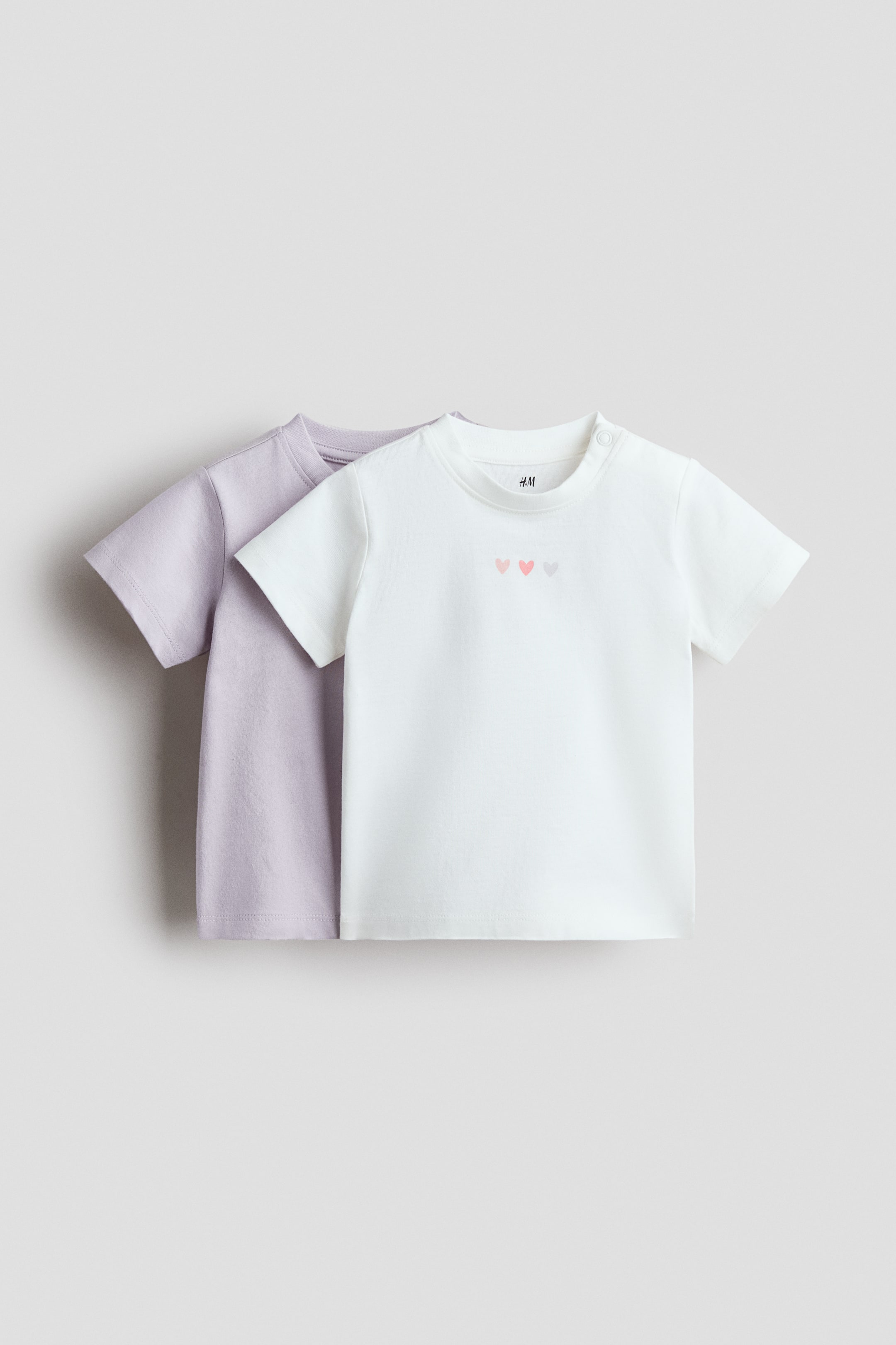 Agrandir l'image: Un lot de 2 T-shirts en coton est présenté, avec un T-shirt blanc superposé devant un T-shirt mauve clair, les deux tournés vers l'avant. Le T-shirt blanc présente un col rond avec deux boutons-pression sur l'épaule gauche et trois petits motifs de cœur en rose, rose clair et gris clair sur la poitrine, tandis que le T-shirt mauve clair affiche un design simple avec un col rond et des manches courtes.