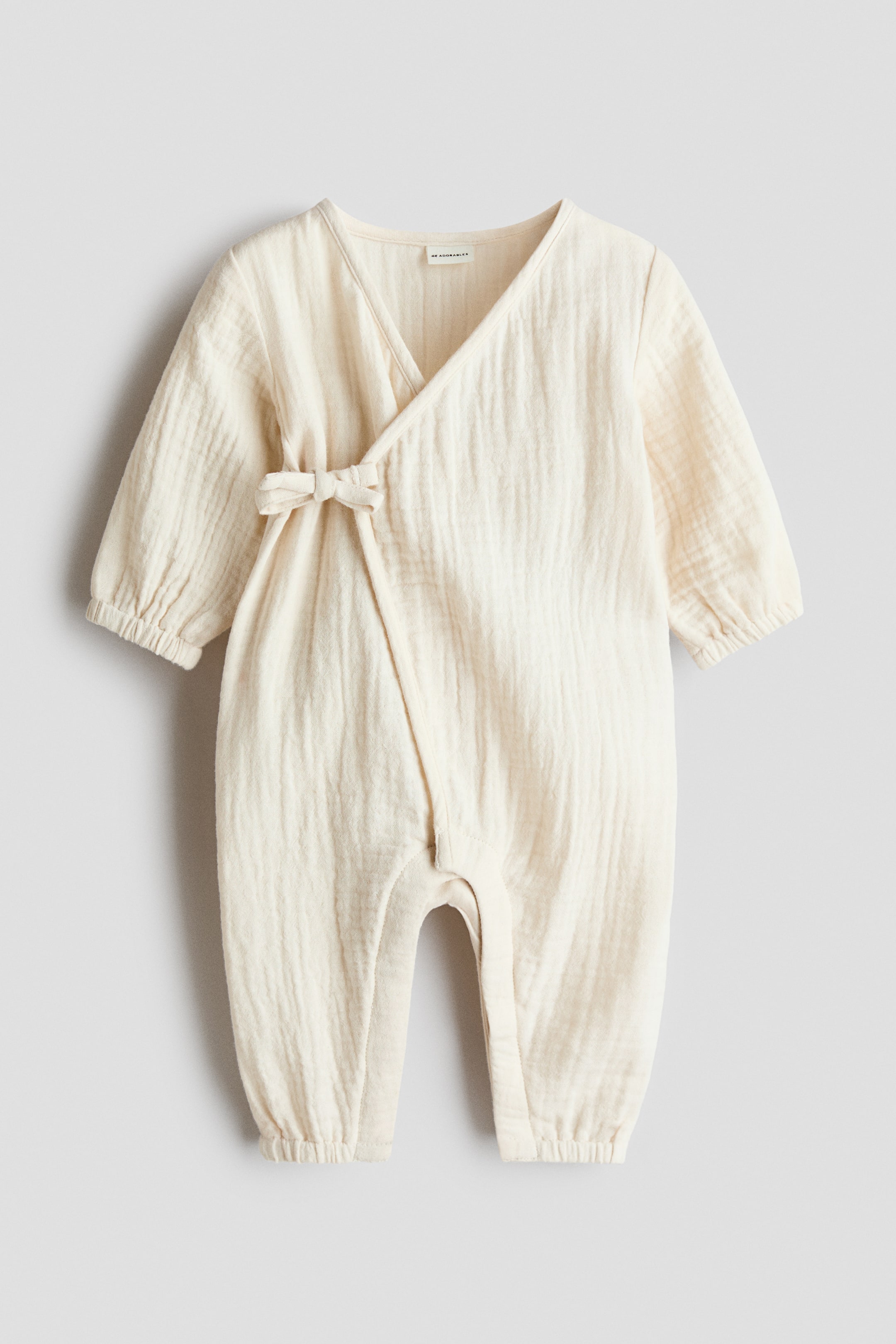 Agrandir l'image: Une barboteuse pour bébé de couleur crème est présentée de face, mettant en valeur un design croisé avec un col en V. Une fermeture à nouer forme un nœud sur le côté gauche, et ses manches longues ainsi que ses jambes se resserrent aux poignets et aux chevilles avec un élastique, tandis que le tissu présente une texture froissée.