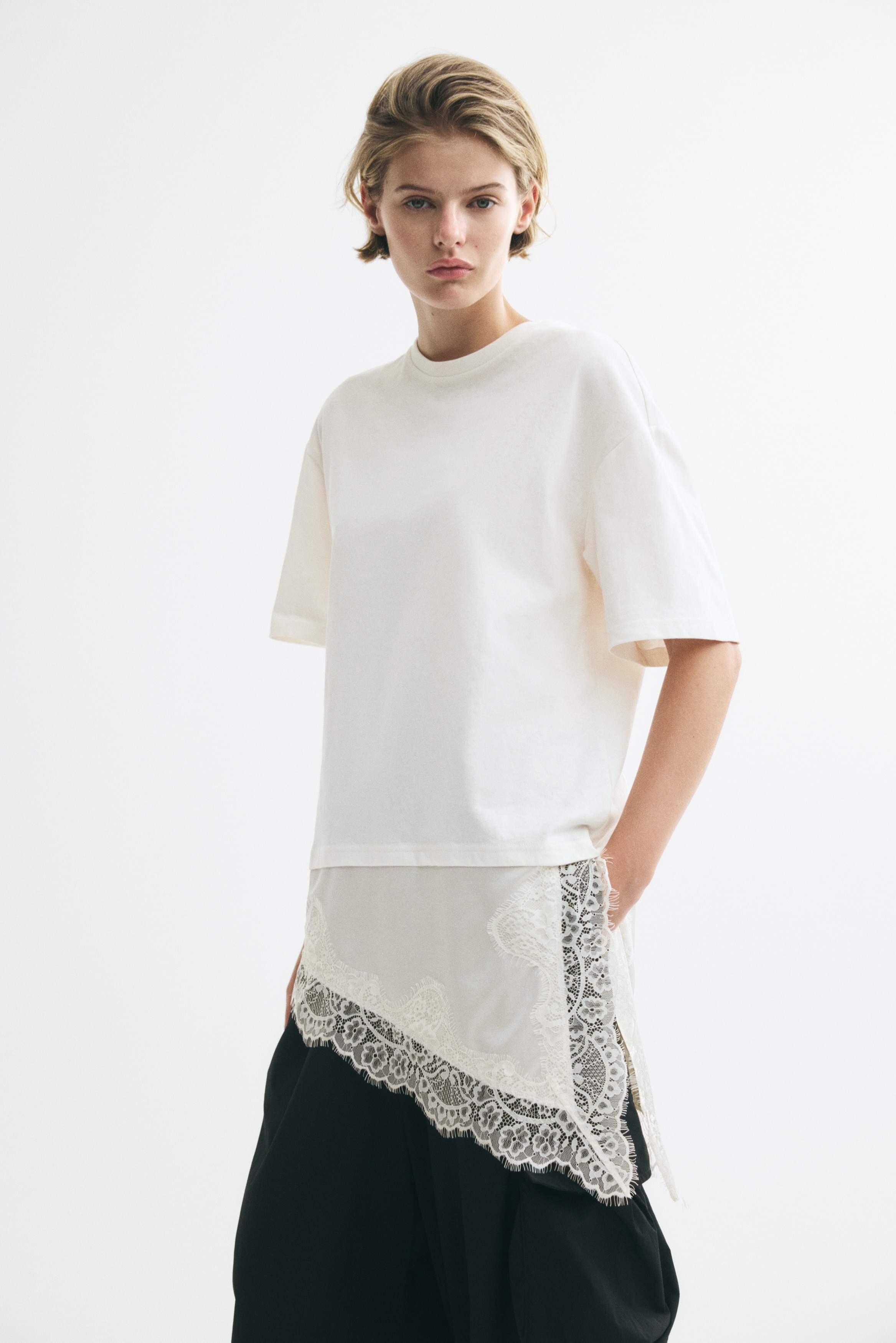 Un t-shirt blanc cassé avec un col rond et des manches courtes et larges, orné d'un panneau de dentelle crème asymétrique superposé sous le tissu principal à l'ourlet. Ce panneau de dentelle s'étend plus longuement sur le côté droit et présente un motif complexe.