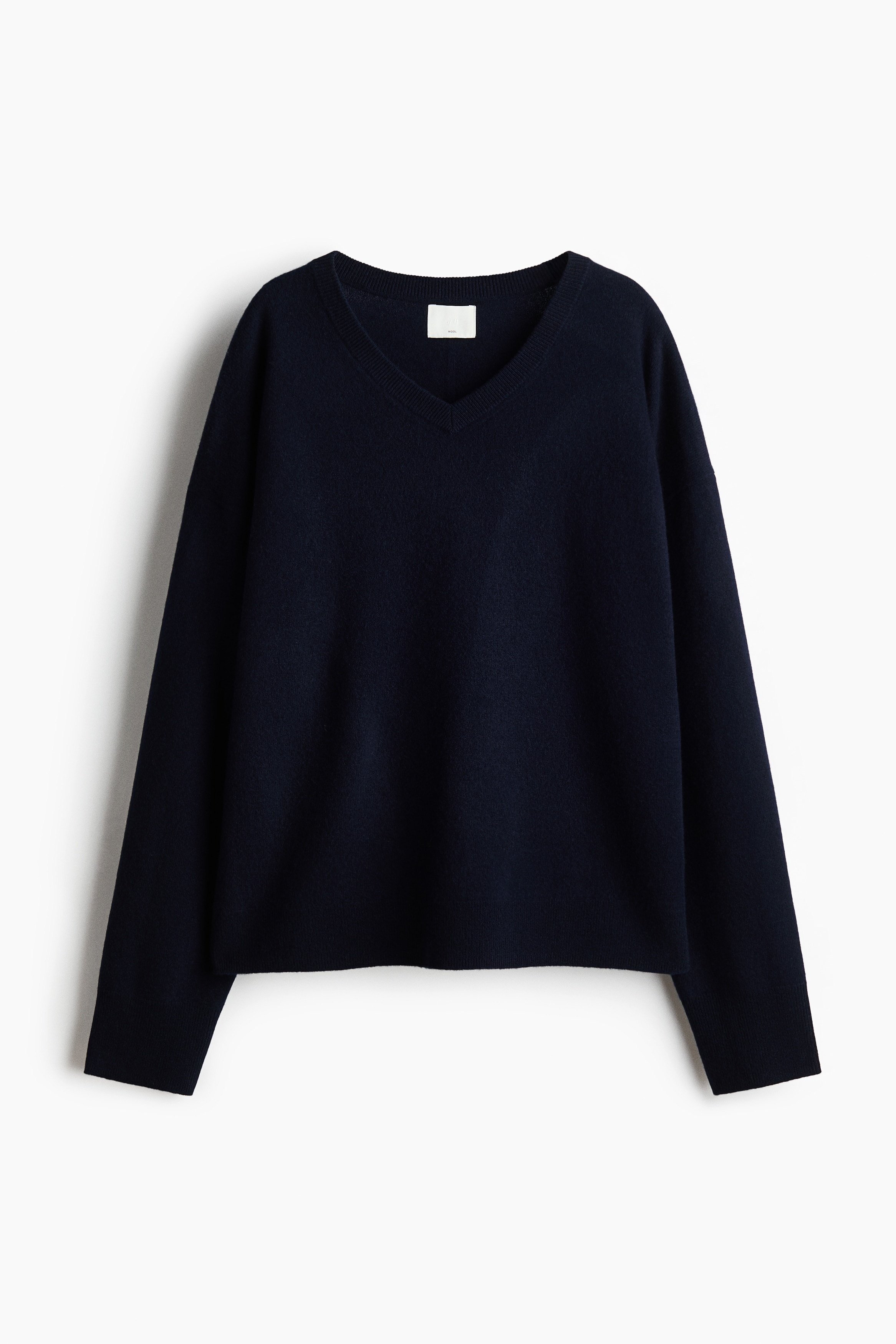 En mÃ¶rk marinblÃ¥ ulltrÃ¶ja i finstickning med ribbad V-ringning, nedhasade axlar och ribbade muddar bÃ¤rs av modellen. Den oversized trÃ¶jan Ã¤r lagerlagd Ã¶ver en vit t-shirt med rund hals och matchas med vita byxor.