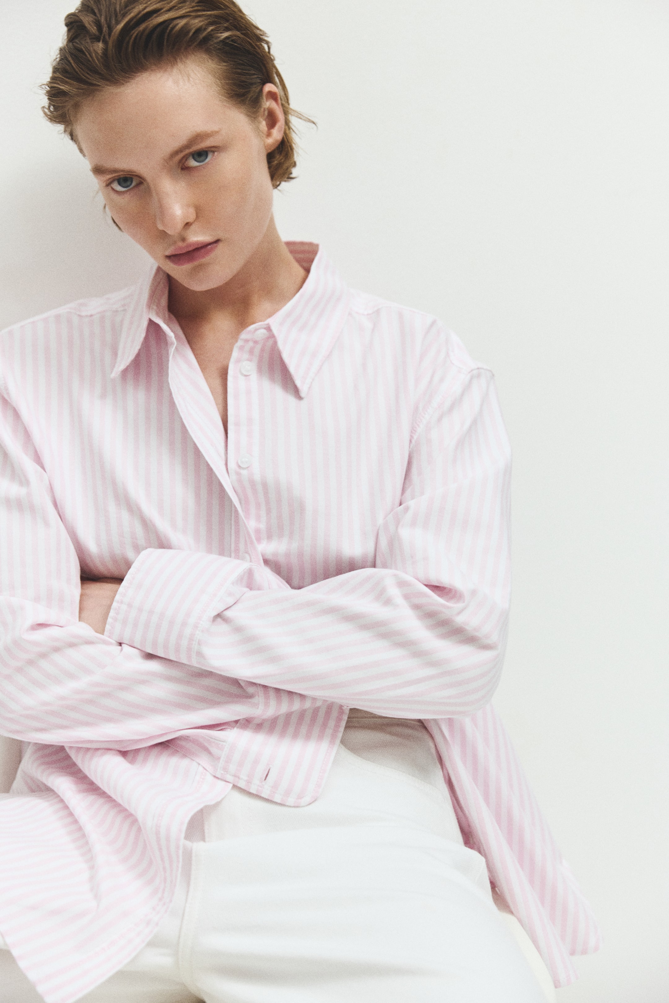 View larger image: The model is wearing a pink and white striped shirt made from washed Oxford cotton. This relaxed-fit shirt includes a collar, several open buttons down the front, dropped shoulders, long sleeves with buttoned cuffs, and a rounded hem. The model has their arms crossed, wearing the shirt untucked over white pants.