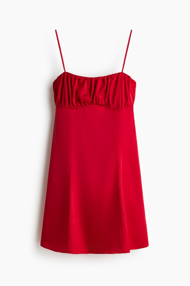A red mini dress is shown facing front, featuring thin spaghetti straps and a gathered, ruched bust with a curved underbust seam. The smooth fabric is draping into a slightly flared A-line skirt with subtle vertical panel seams. - 1