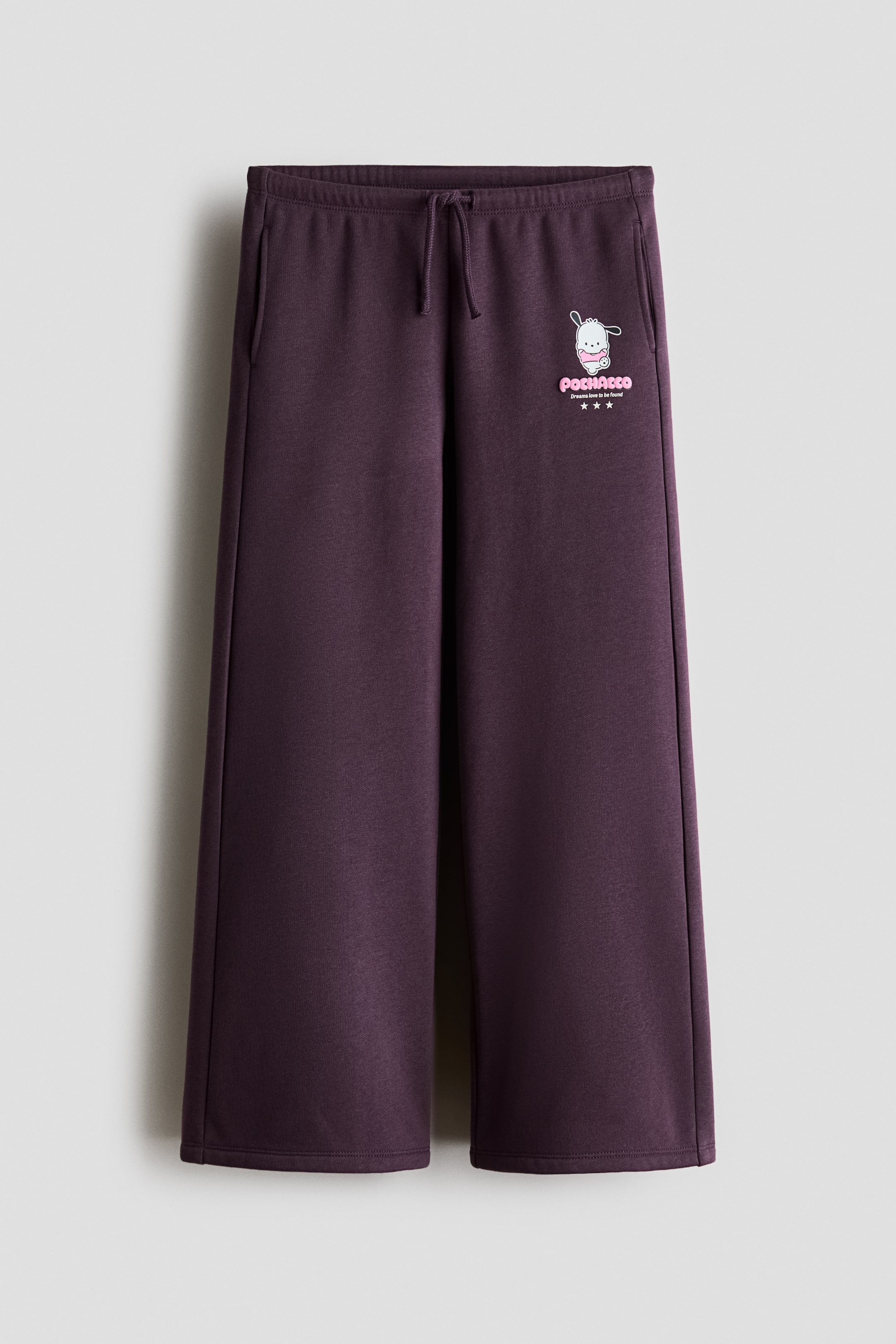 View larger image: Dark purple wide-leg sweatpants are shown from the front, featuring an elasticated drawstring waistband and two side pockets. A graphic of a white cartoon character with pink accents, "POCHACCO Dreams love to be found" text, and three pink stars is printed on the upper left leg.
