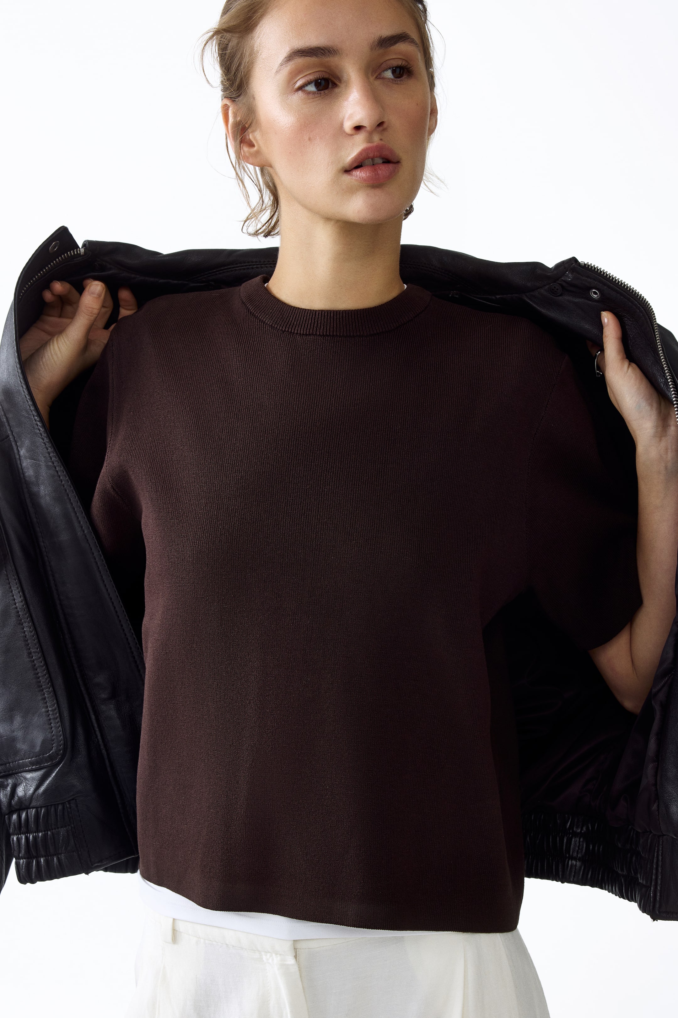 View larger image: A dark brown knit short-sleeve top is being worn, featuring a ribbed crew neck and fine vertical ribbing throughout the fabric. A white layer is visible beneath its straight hem.