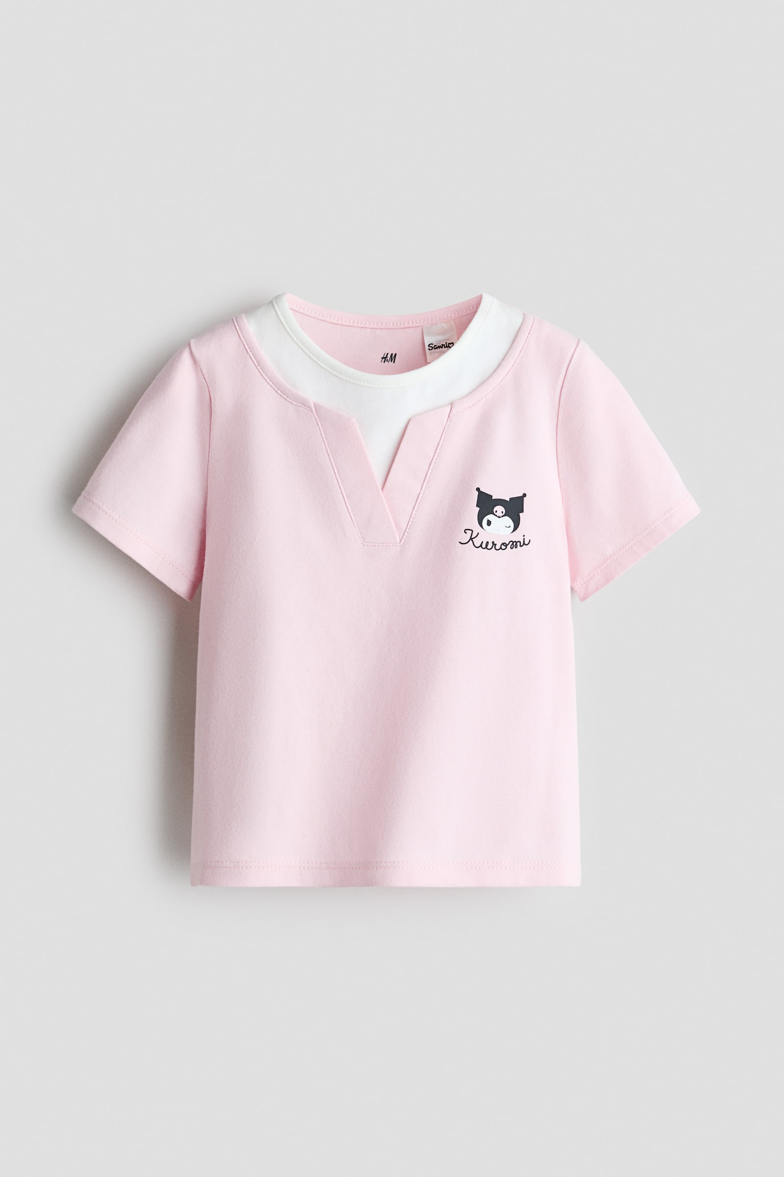 A light pink, short-sleeved T-shirt is shown from the front, featuring a V-shaped neckline that reveals a layered white crew-neck insert underneath. The chest features a small, black-and-white graphic print of the character Kuromi, with the name written in cursive below it.