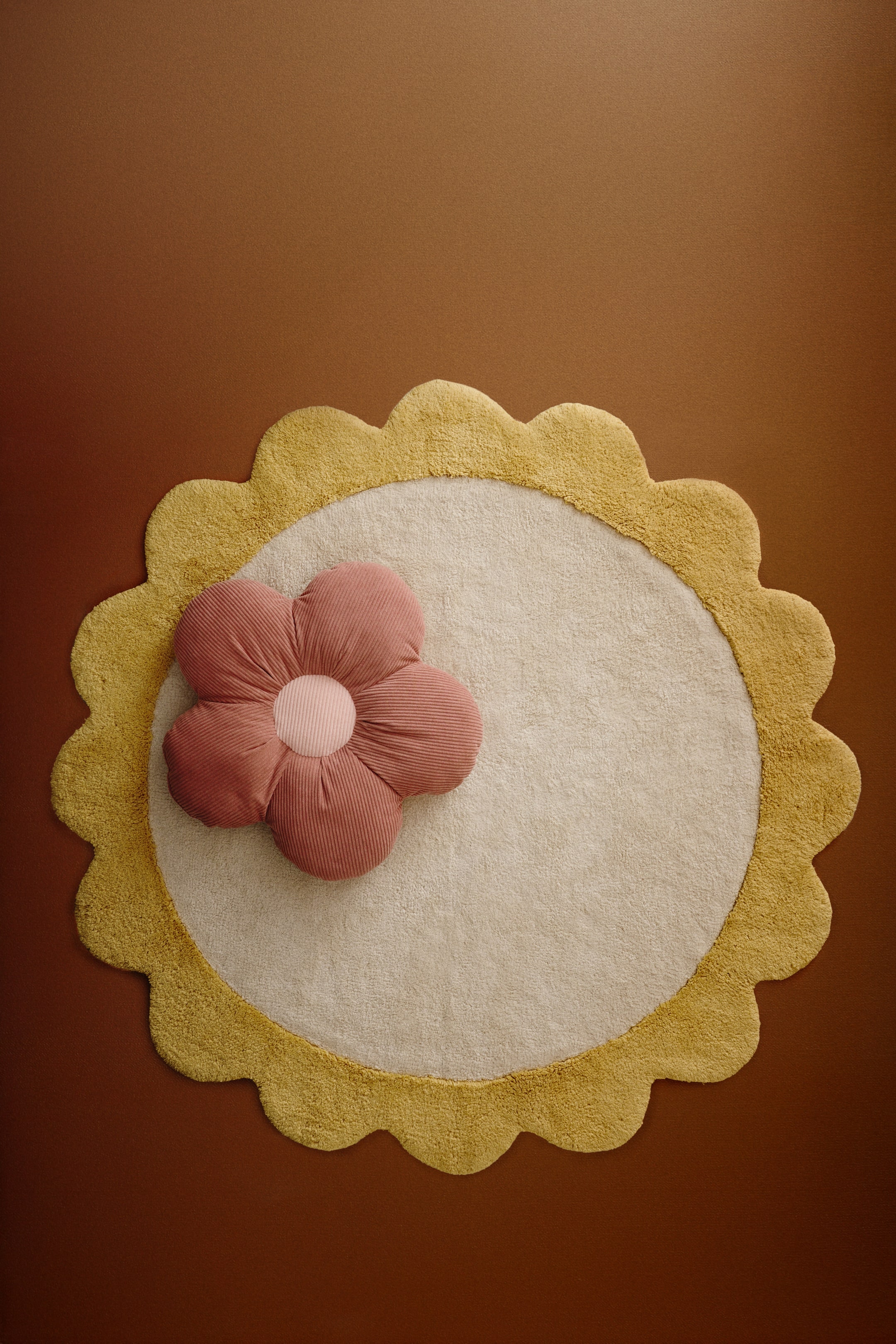 Nagyobb kép megtekintése: "Egy kerek krémszínű szőnyeg sárga hullámos szegéllyel fekszik síkban. Egy rózsaszín virág alakú párna bordázott textúrával és világosabb rózsaszín középpel pihen a szőnyeg felszínén."