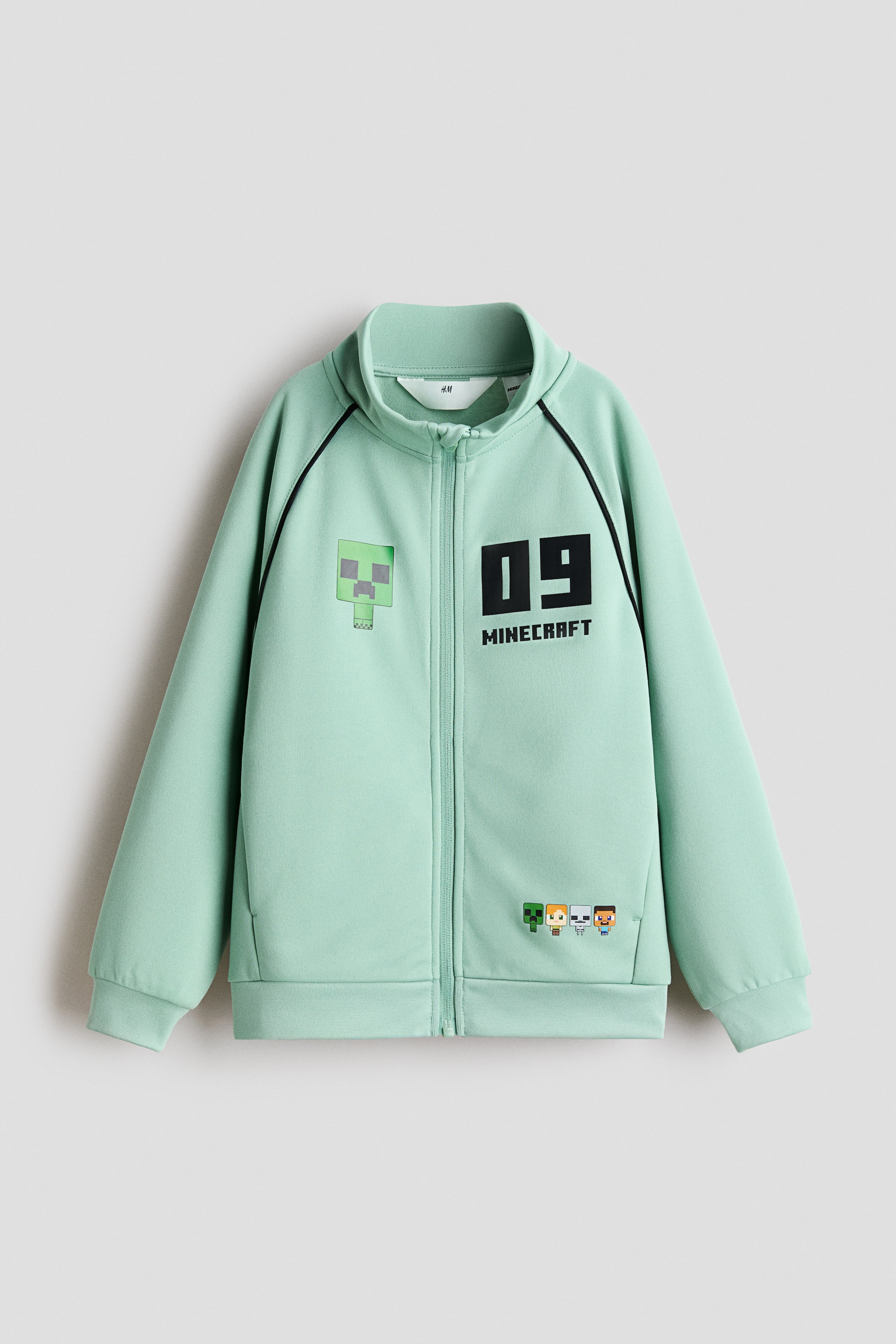 Grösseres Bild ansehen: Die Jacke ist VON VORNE zu sehen und besteht aus einem mintgrünen, schweren Jersey mit leichtem Glanz. Sie verfügt über einen Stehkragen, einen Reissverschluss vorne mit scheuerschützendem Kinnschutz, lange Raglanärmel, unauffällige Seitentaschen sowie Rippbündchen an Ärmelabschlüssen und Saum. Auf der Vorderseite befinden sich Print-Motive, darunter eine verpixelte grüne Figur auf der linken Brust, der Schriftzug „09 MINECRAFT“ auf der rechten Brust sowie kleine verpixelte Figuren in Saumnähe.