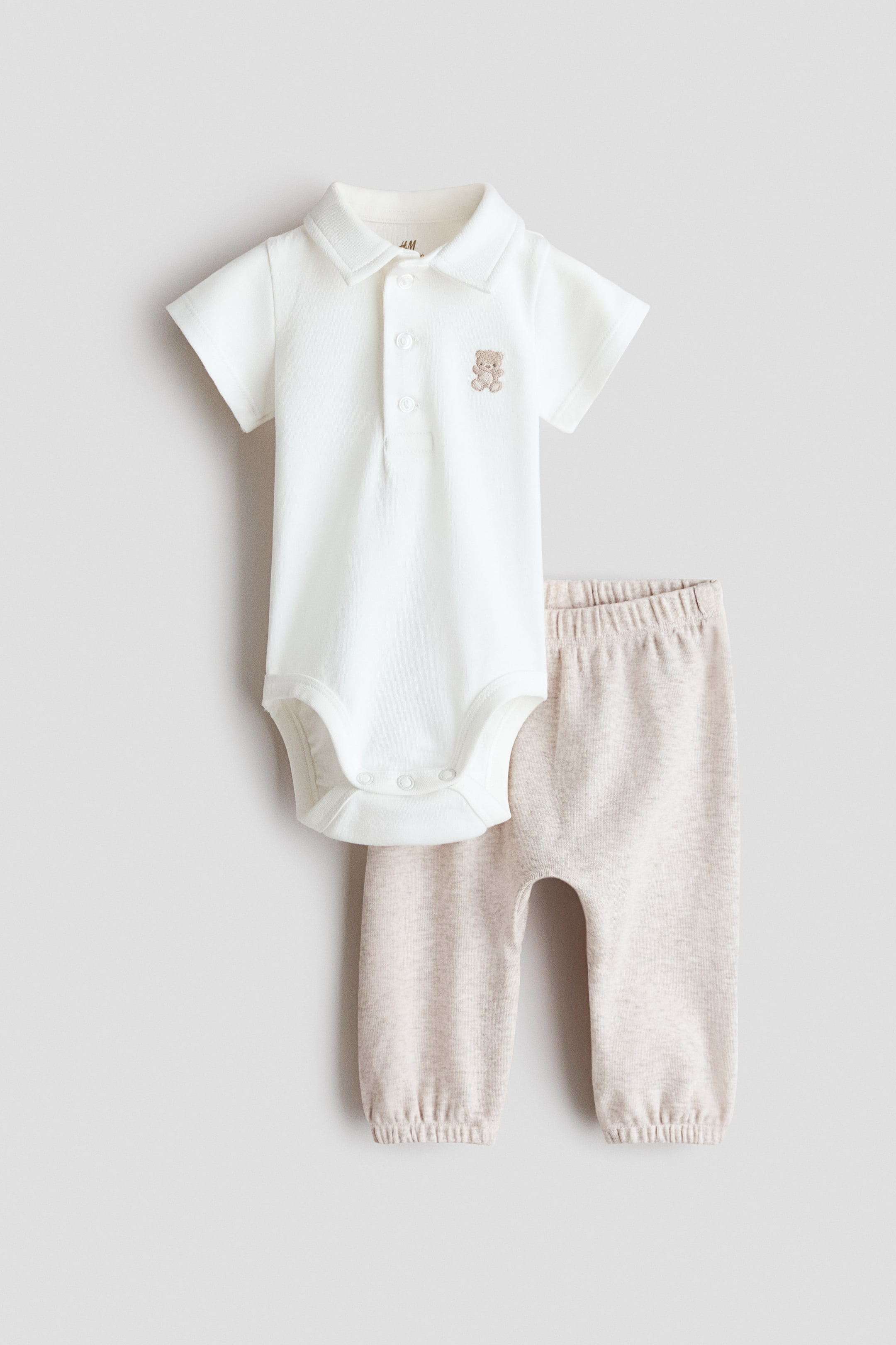View larger image: Introducing a baby two-piece set, featuring a white short-sleeved polo bodysuit with a collar, a three-button placket, a small embroidered brown teddy bear on the left chest, and snap fasteners at the crotch. It is paired with light beige pants that come with an elasticated waistband and elasticated cuffs at the ankles.