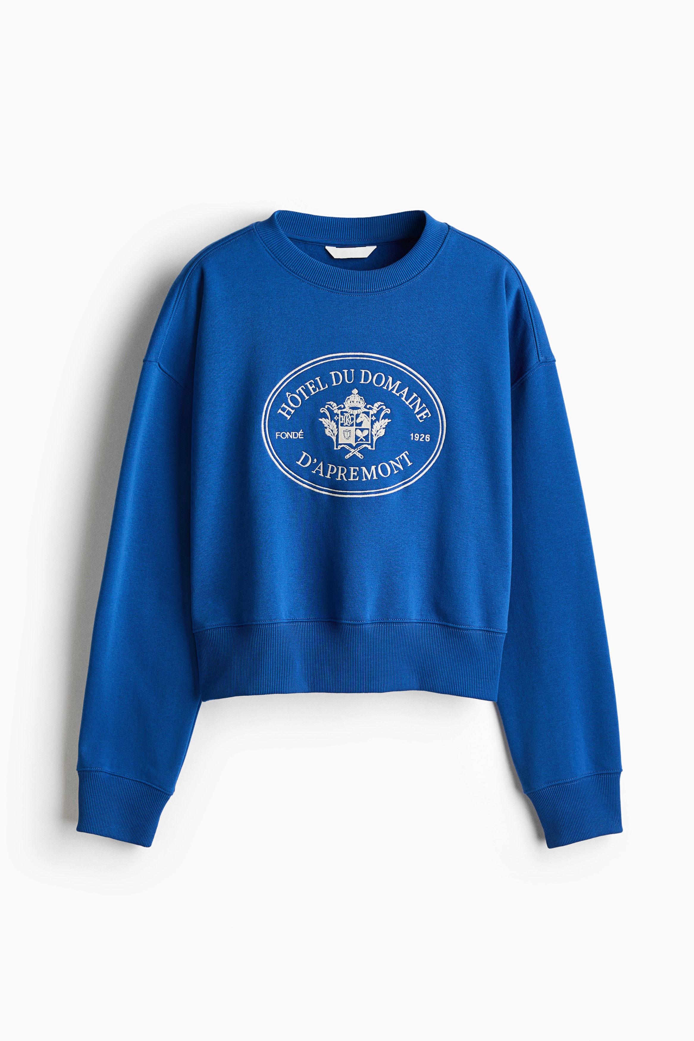 A royal blue sweatshirt featuring a white circular crest graphic printed on the chest is being worn by the model. The model is raising both hands to their head, highlighting the sweatshirt's ribbed crew neckline and cuffs.