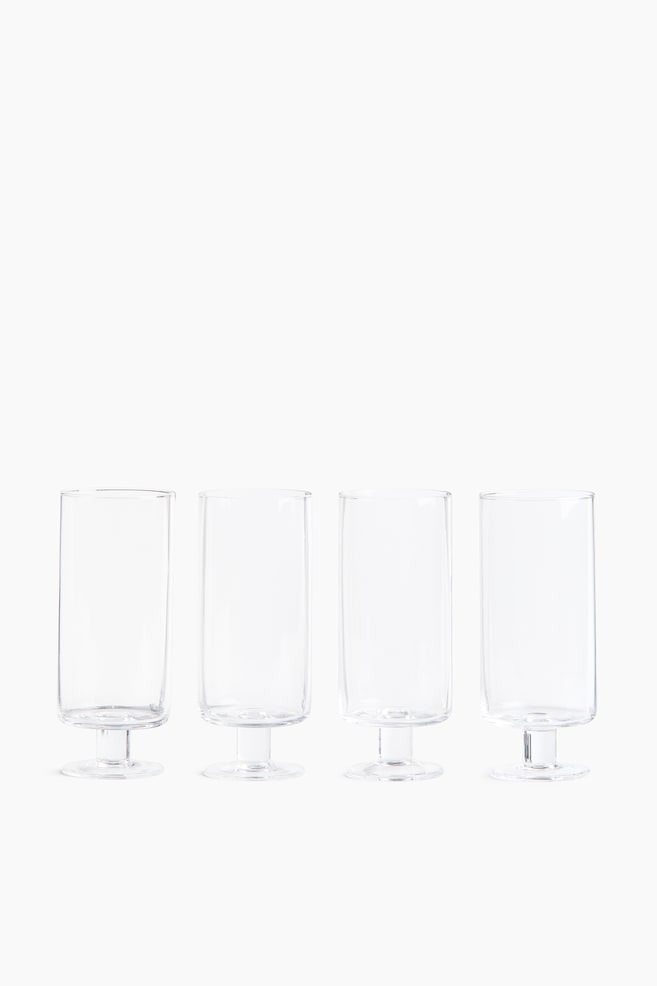 Four mouth-blown, footed highball glasses with a straight-sided design are arranged in a horizontal row, facing forward. Each clear glass rests on a short, thick stem and a flat, circular base, highlighting its transparent, cylindrical form. - 1