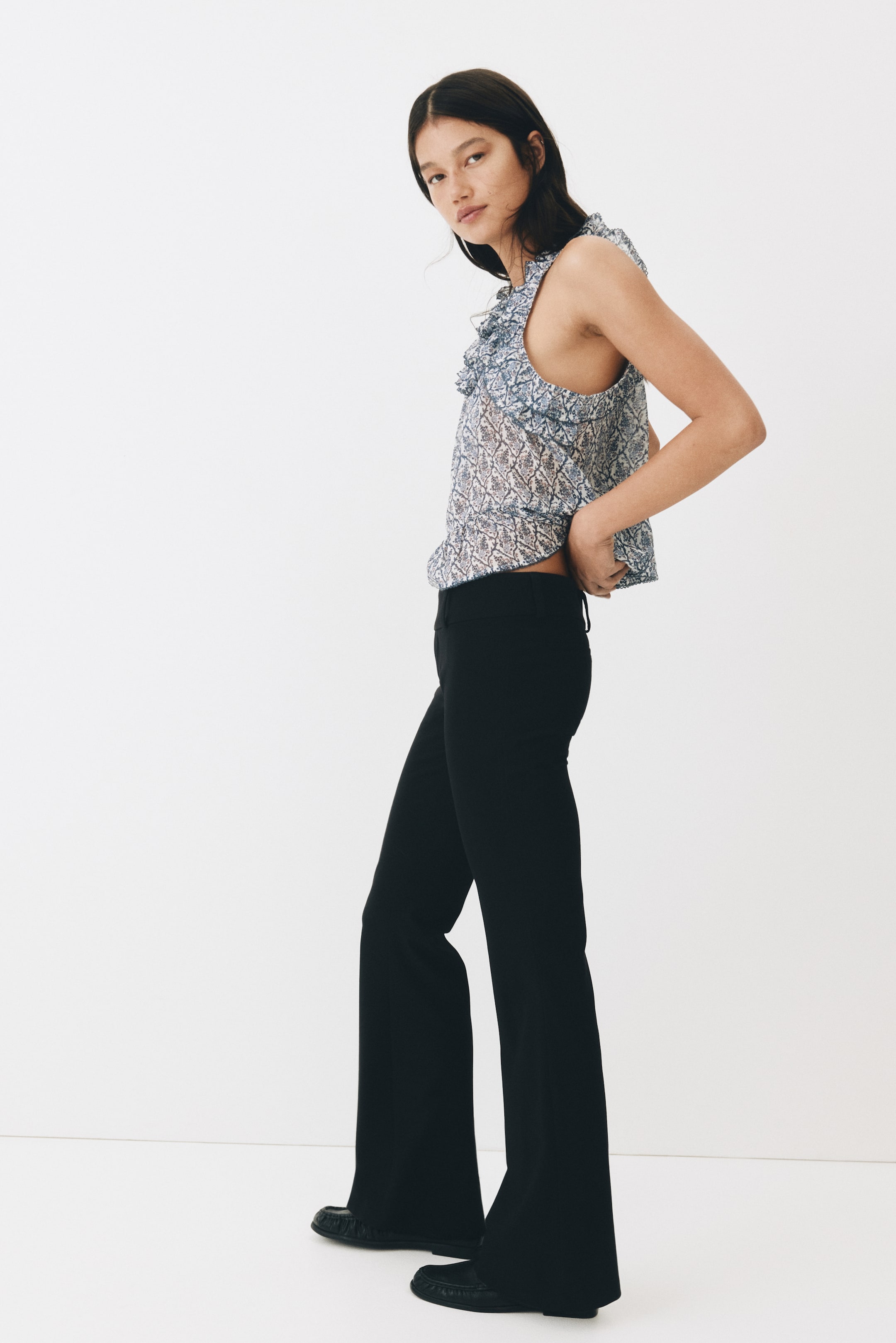 View larger image: The model is showcasing fitted, tailored black trousers featuring a low waist and flared legs. She's adjusting her top, subtly revealing the low waistband of the trousers.