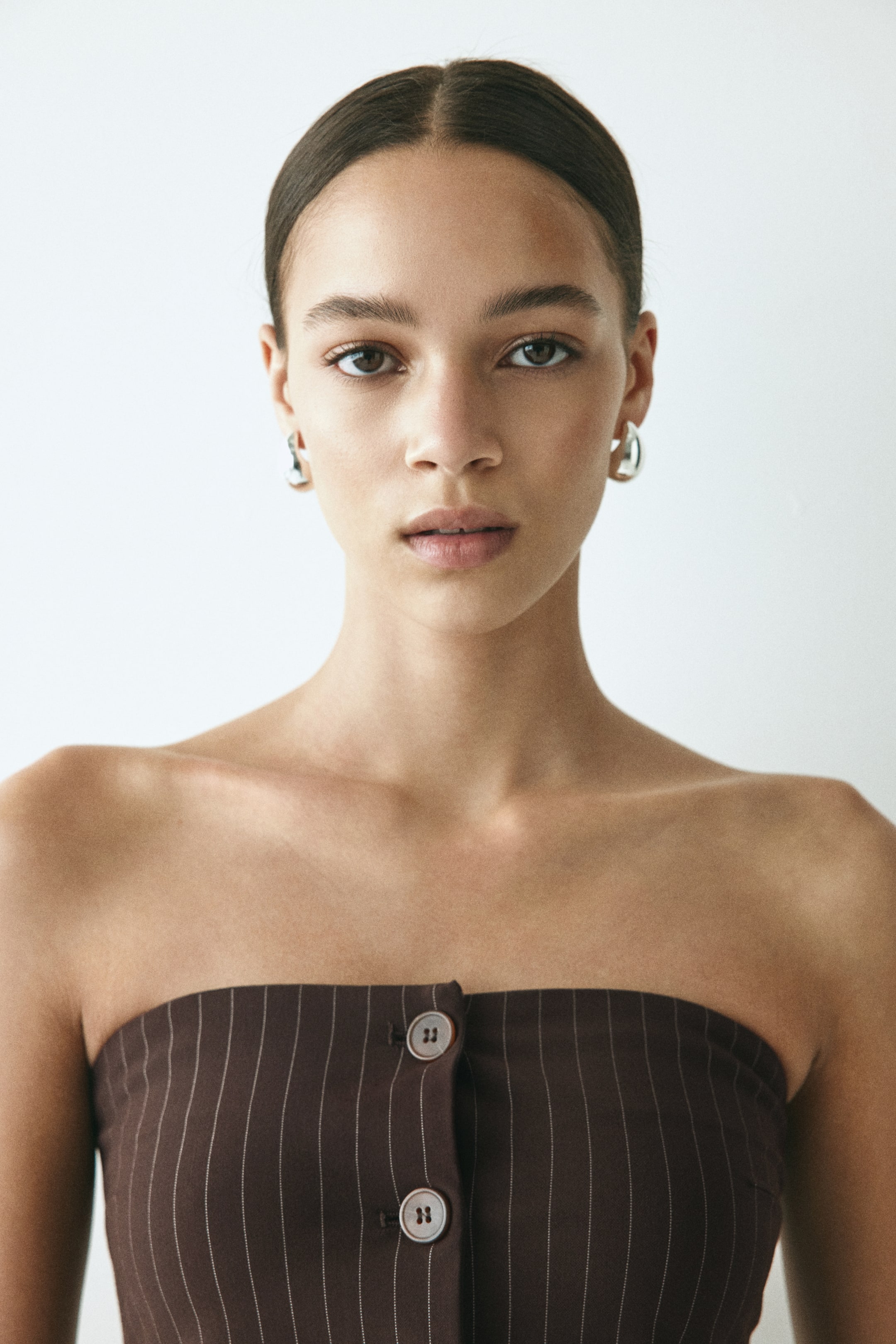 Ver imagem maior: Uma peça castanho-escuro sem alças apresenta finas riscas verticais em tom claro. Dois botões redondos, de cor clara, são visíveis na sua parte frontal.