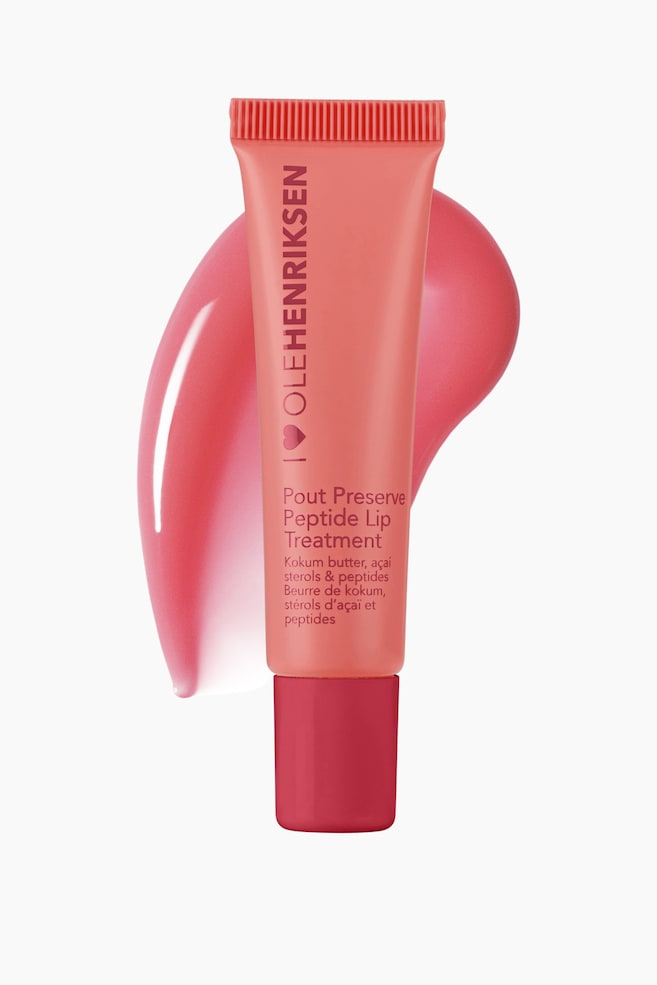 Et ferskenrosa leppebehandlingsrør står oppreist, vendt fremover, med teksten "OLEHENRIKSEN" og "Pout Preserve Peptide Lip Treatment", og er toppet med en mørkere rosa base. En blank, ferskenrosa strek av produktet vises bak og delvis under røret. - 1
