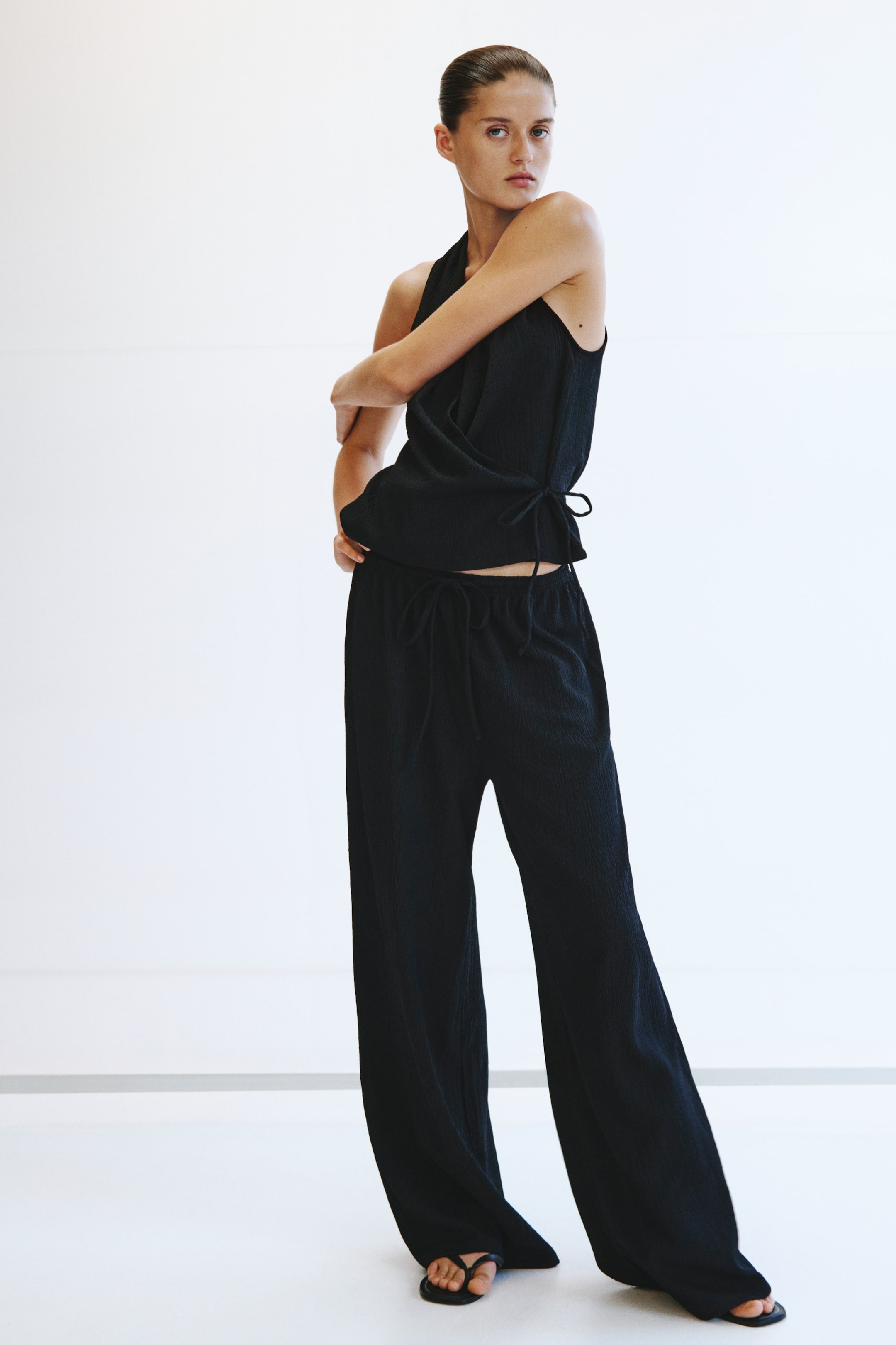 Agrandir l'image: Un haut sans manches noir froissé avec un devant style cache-cœur et une fermeture à nouer sur le côté est porté avec un pantalon large noir froissé assorti, doté d'une taille à cordon de serrage.