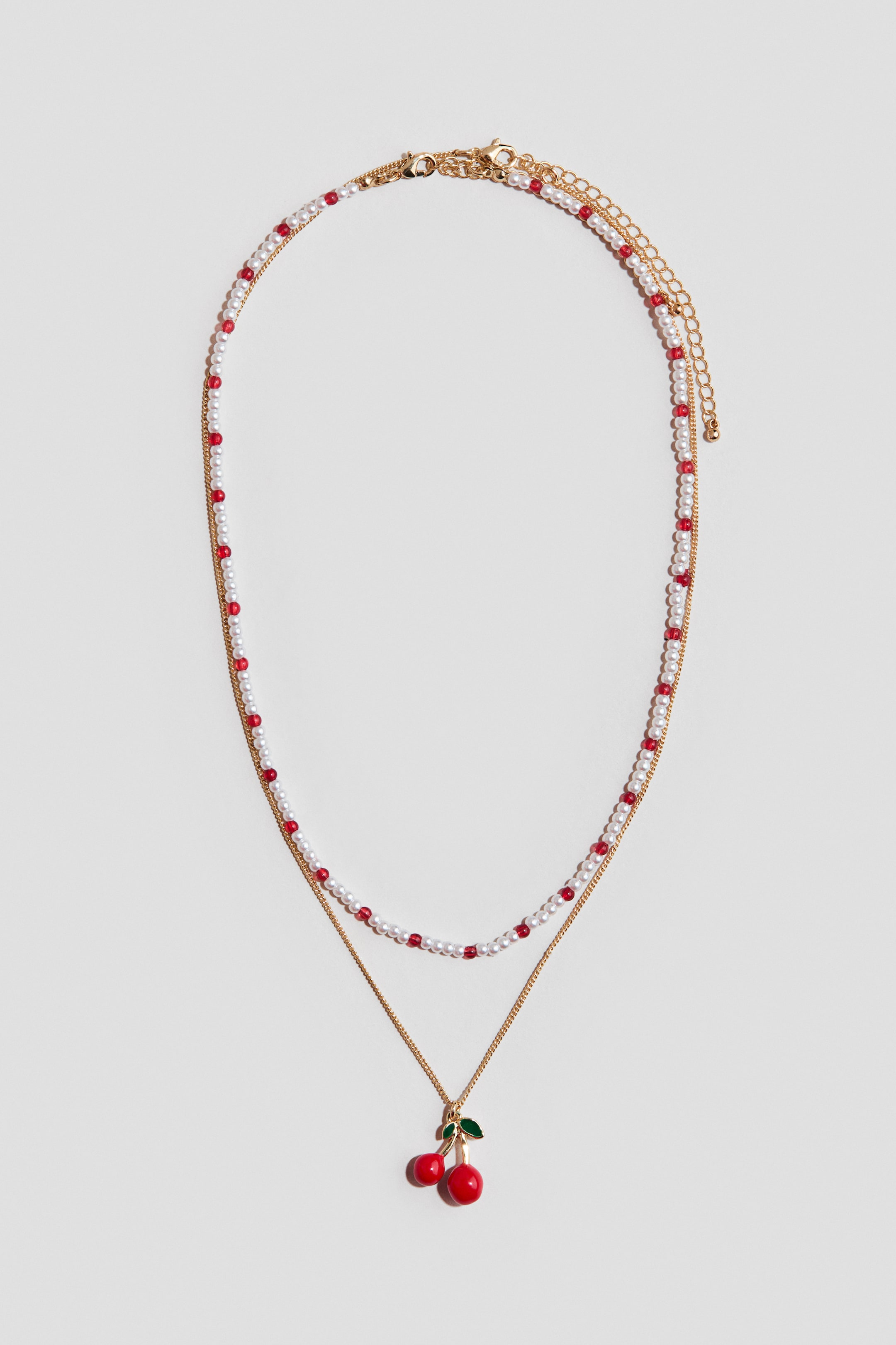 Se større billede: Et lagdelt halskæde viser to tråde: den øverste tråd har små hvide perler, der skifter med røde perler, og den nederste tråd består af en guldfarvet kæde, hvorfra et rødt kirsebærvedhæng med grønne blade hænger. Guldfarvede hummerlåse og en forlængerkæde er synlige bag på halskæden.