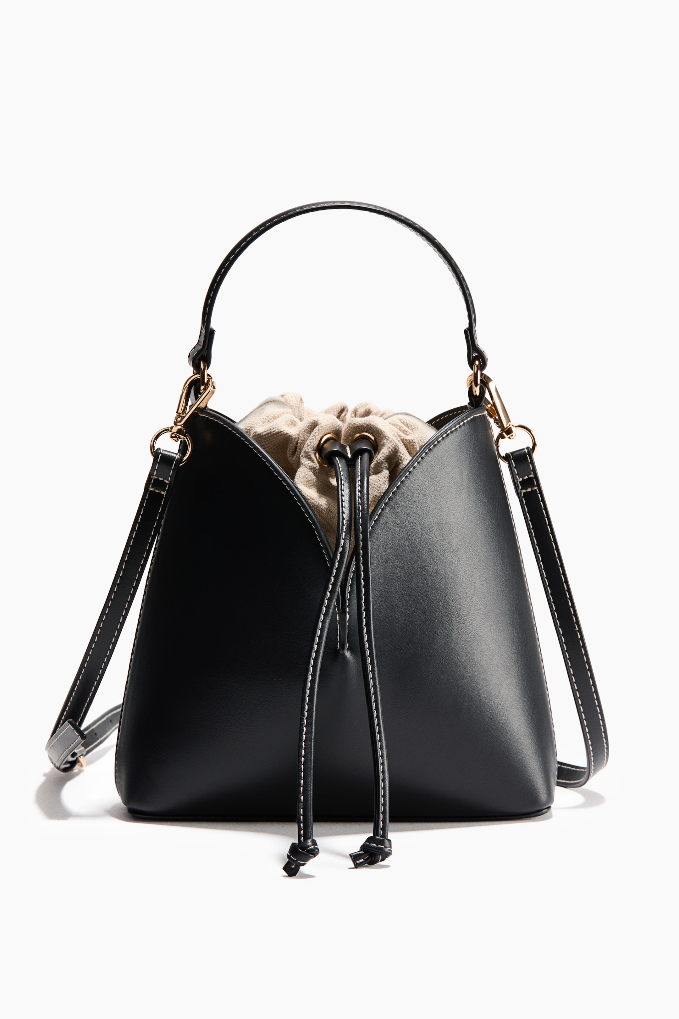 View larger image: A black bucket bag is presented front, featuring a tapered body with white contrast stitching. A light beige drawstring inner pouch, secured with gold-toned eyelets and black drawstrings, is visible at the top. The bag includes a curved top handle and a detachable shoulder strap with white contrast stitching.