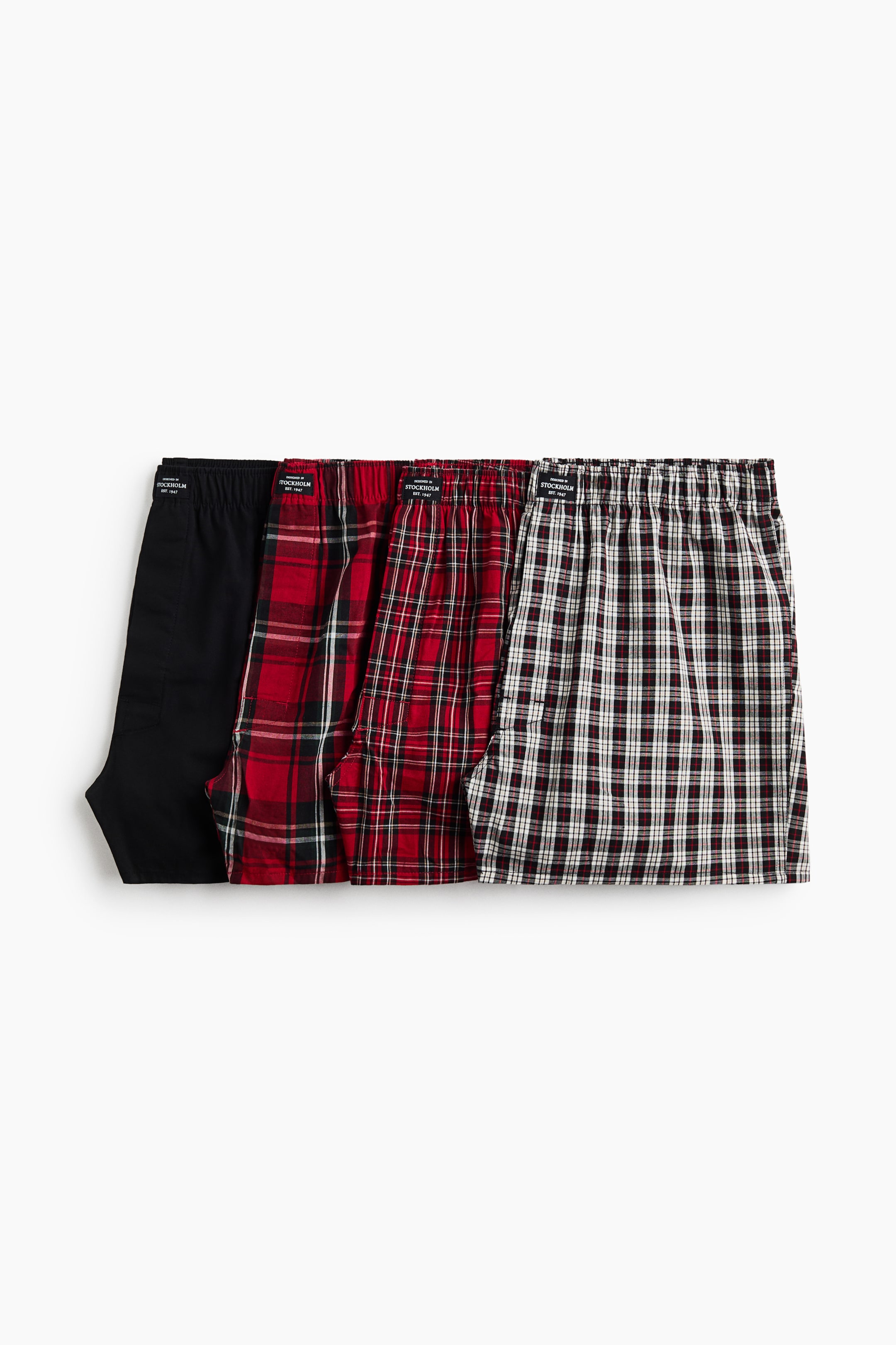 View larger image: Four pairs of woven boxer shorts are laid flat in an overlapping row, showing a solid black pair, two red and black plaid variations, and one black and white plaid pair. Each pair features an elasticised waistband with a small centre-front label and relaxed, long legs.