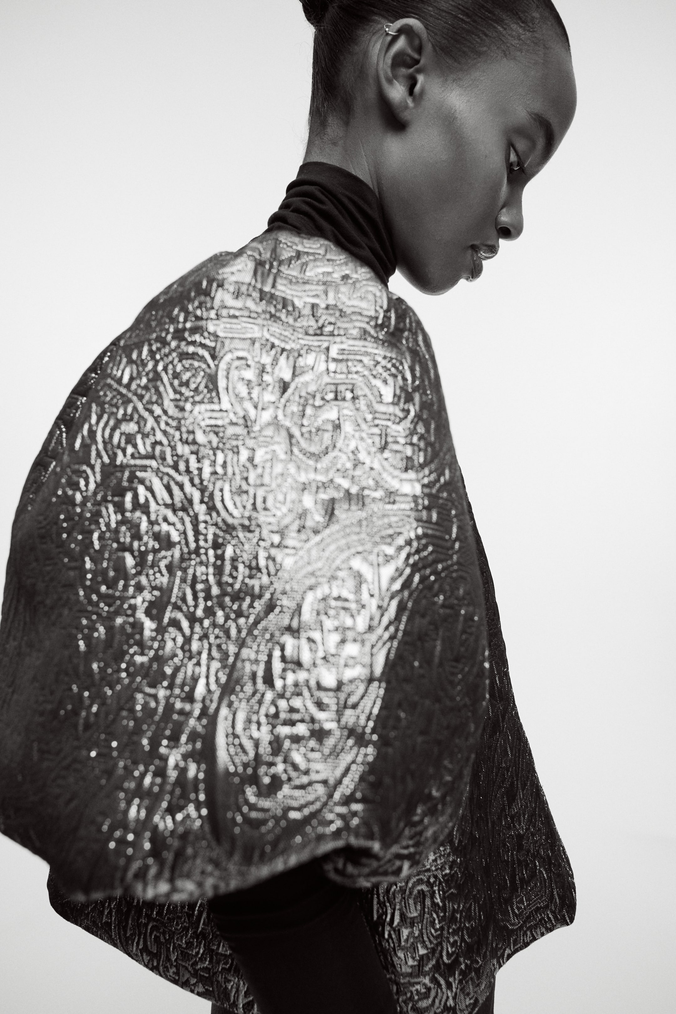 Zobacz większy obraz: Obszerna odzież wierzchnia, prawdopodobnie kurtka lub peleryna, jest prezentowana z boczno-tylnego kąta. Ma skomplikowany, wypukły wzór o metalicznym wyglądzie na swojej powierzchni i jest noszona na czarnym golfie.