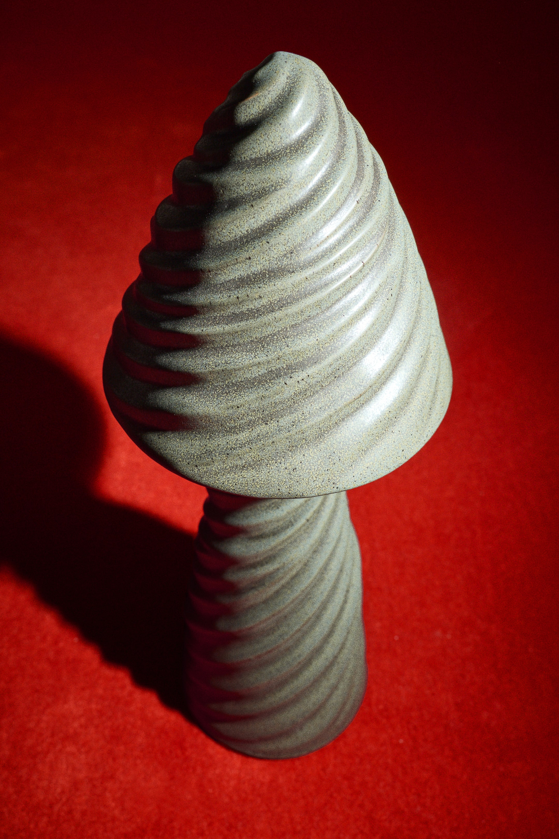 Agrandir l'image: Une lampe de table en grès est posée debout, mettant en valeur un design sculptural en forme de champignon, avec des reliefs doux et ondulés enveloppant son sommet conique et sa base cylindrique. La lampe présente une finition émaillée réactive gris-vert mouchetée et texturée.