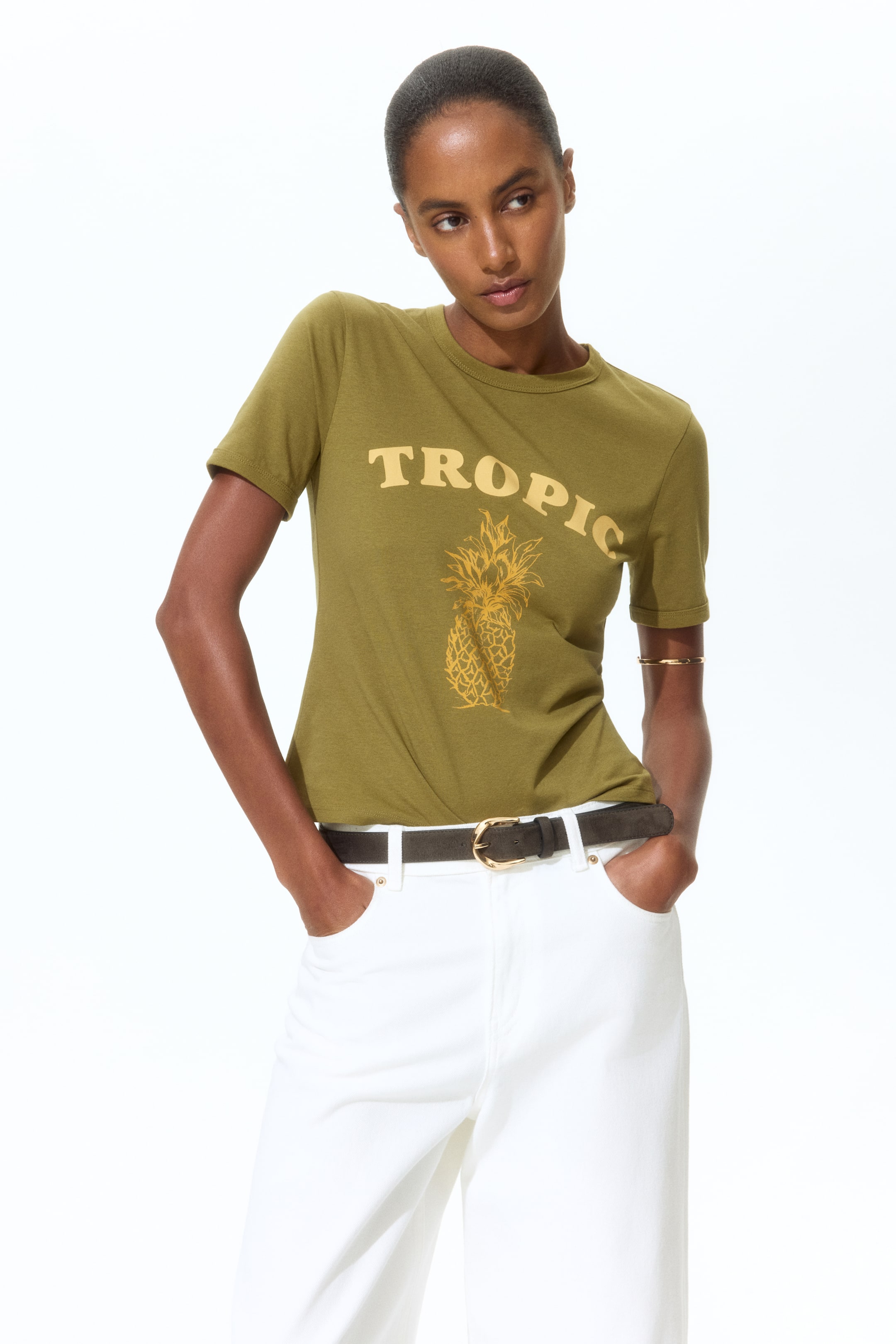 View larger image: An olive green short-sleeve t-shirt showcases a yellow "TROPIC" text print above a yellow pineapple illustration on the front. It's styled tucked into white trousers.