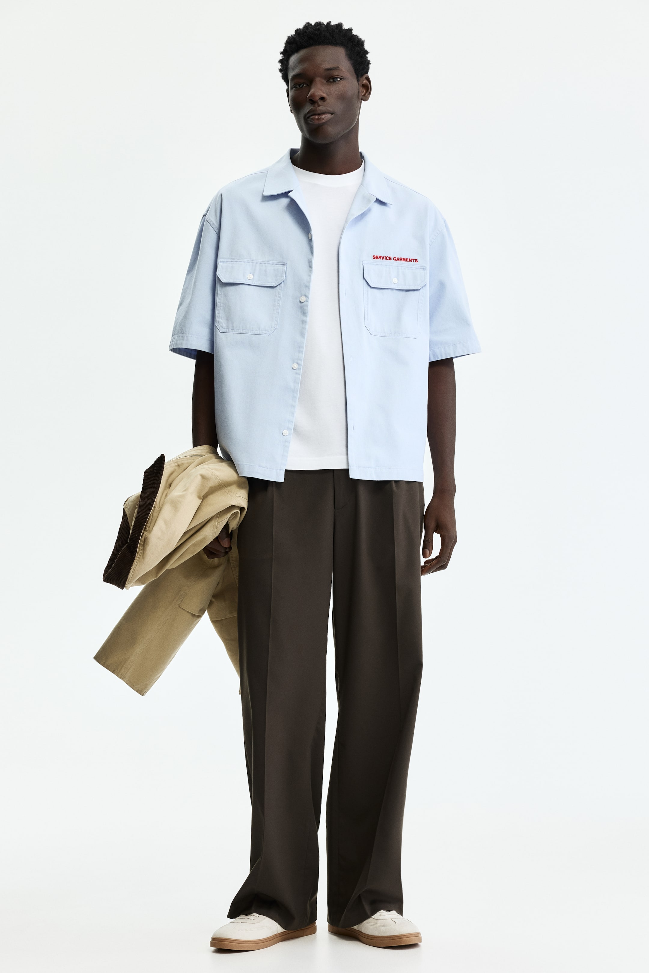 View larger image: A light blue short-sleeved shirt is worn open over a white t-shirt, showcasing a camp collar and two chest patch pockets with flap and button closures. Red embroidered text "SERVICE GARMENTS" appears above the left chest pocket, and white buttons line the front placket.