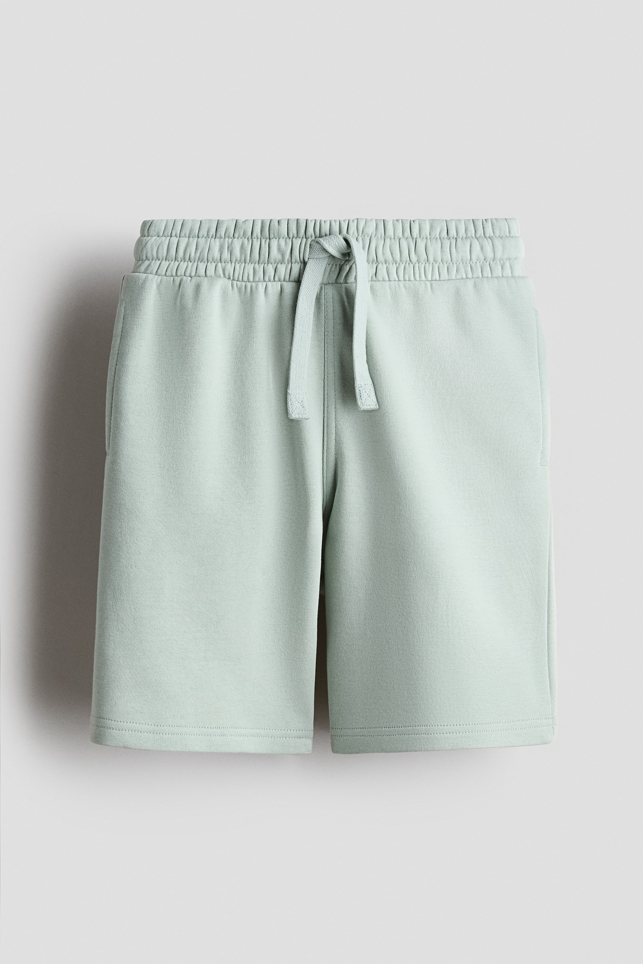 View larger image: Light green knee-length shorts crafted from sweatshirt fabric, showcasing an elasticated drawstring waist with the drawstring tied at the front. Discreet welt pockets are visible along the side seams, slightly positioned towards the front.