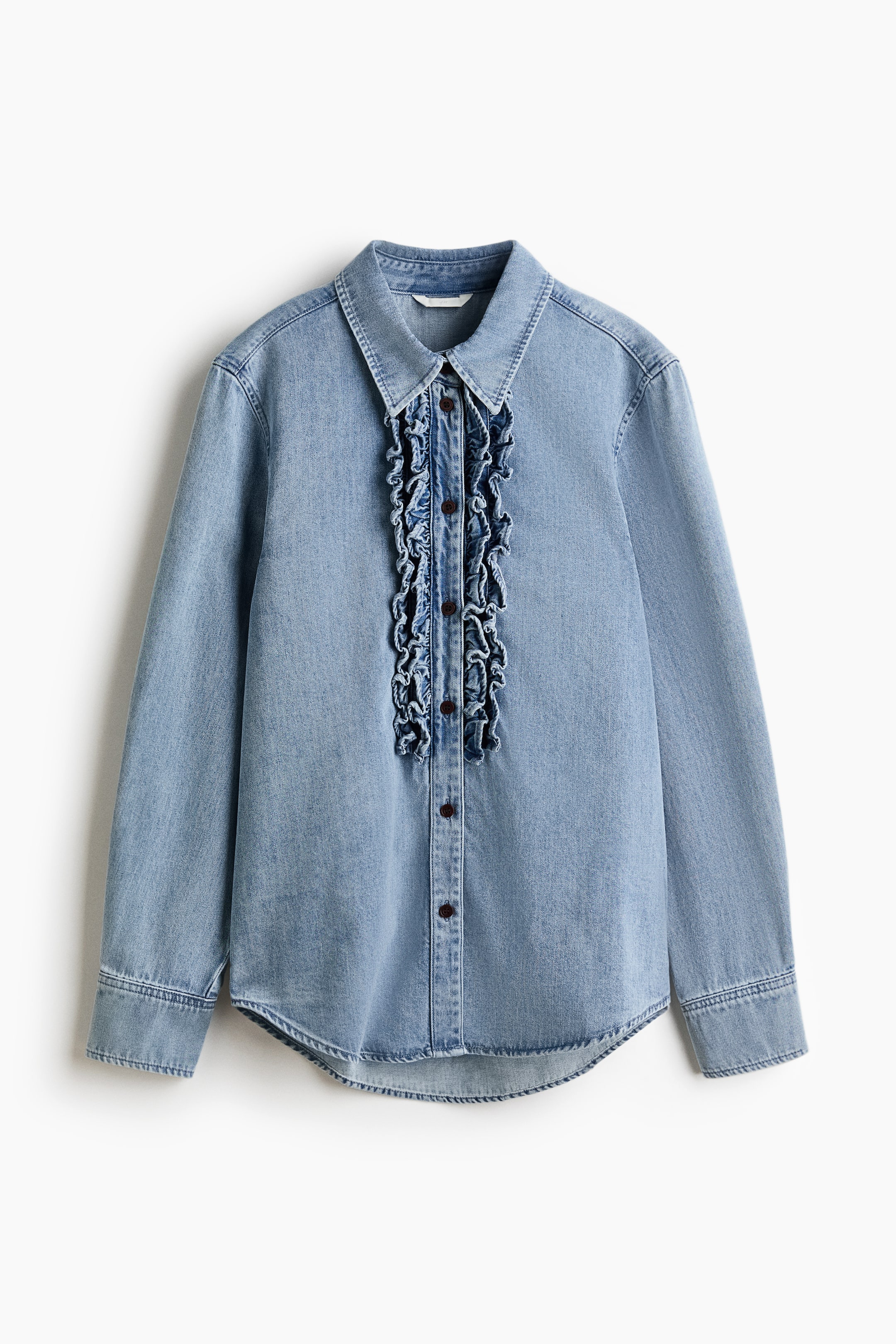 View larger image: A light blue denim shirt is shown from the front, featuring a classic pointed collar and long sleeves with buttoned cuffs. A central placket with dark-coloured buttons is adorned with vertical ruffles on both sides, and the hemline is curved.