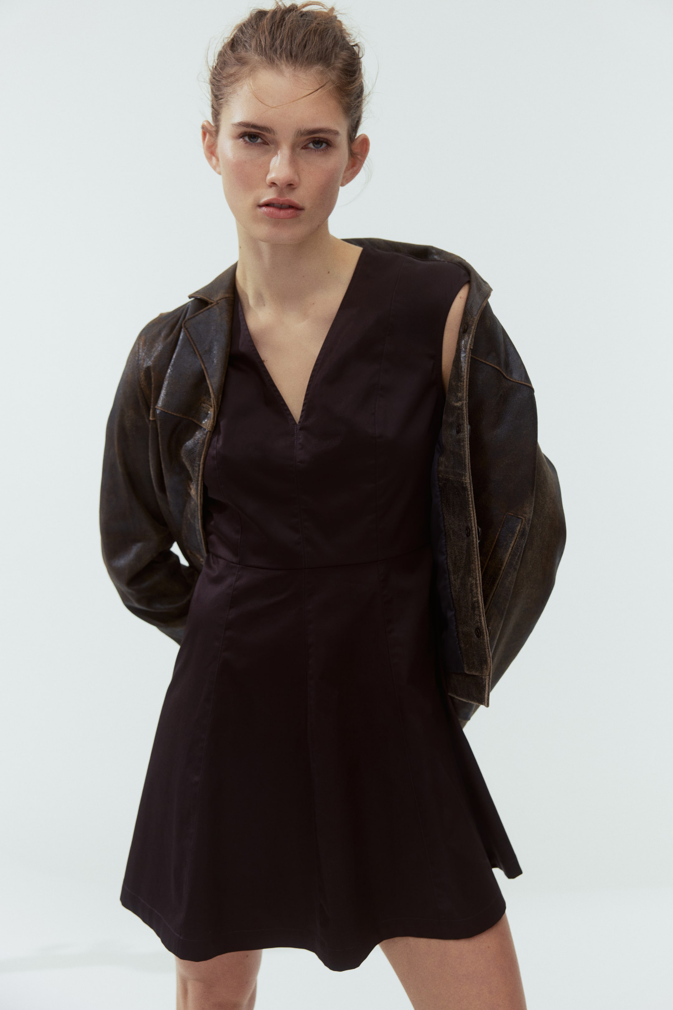 Ampliar la imagen: Un vestido oscuro sin mangas con escote en V presenta un corpiño entallado con líneas de costura visibles y una falda acampanada de corte A. Una chaqueta marrón oscuro con acabado desgastado está colocada sobre sus hombros, luciendo un cuello clásico.