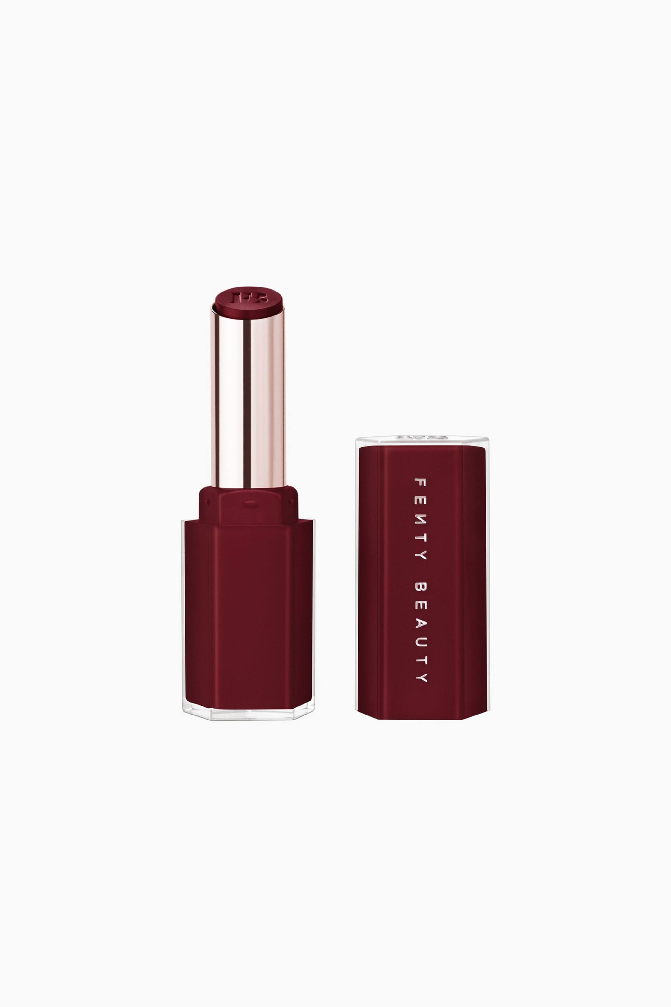 Näytä suurempi kuva: Gloss Bomb Stix High-shine Gloss Stick -kiiltoastipuikko on pystyasennossa eteenpäin suunnattuna irrotetun korkkinsa vieressä. Avoin, syvän viininpunainen, kahdeksankulmainen alaosa pitää sisällään metallinhohtoisen ruusukultaisen sisäosan, jossa tasapäinen kiiltoastipuikko on, ja yhteensopivassa korkissa näkyy pystysuuntainen "FENTY BEAUTY" -logo.