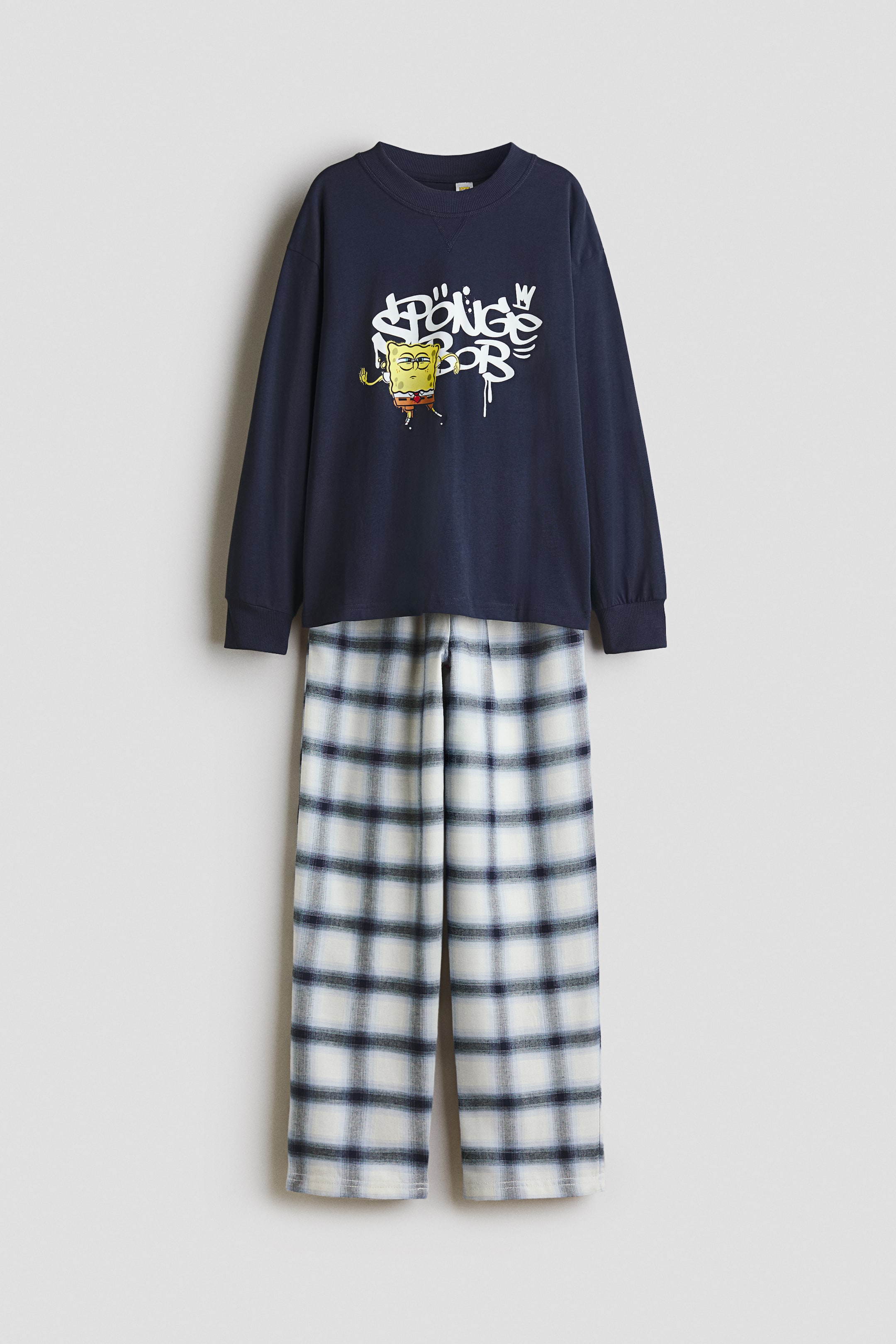 Grotere afbeelding bekijken: Een tweedelig pyjama-set wordt frontaal gepresenteerd. De marineblauwe, lange mouwentop heeft een witte graffiti-stijl "SpongeBob"-afbeelding met het cartoonfiguur op de borst, samen met een ronde hals en geribbelde boorden. De bijpassende lange broek toont een wit en blauw geruit patroon.