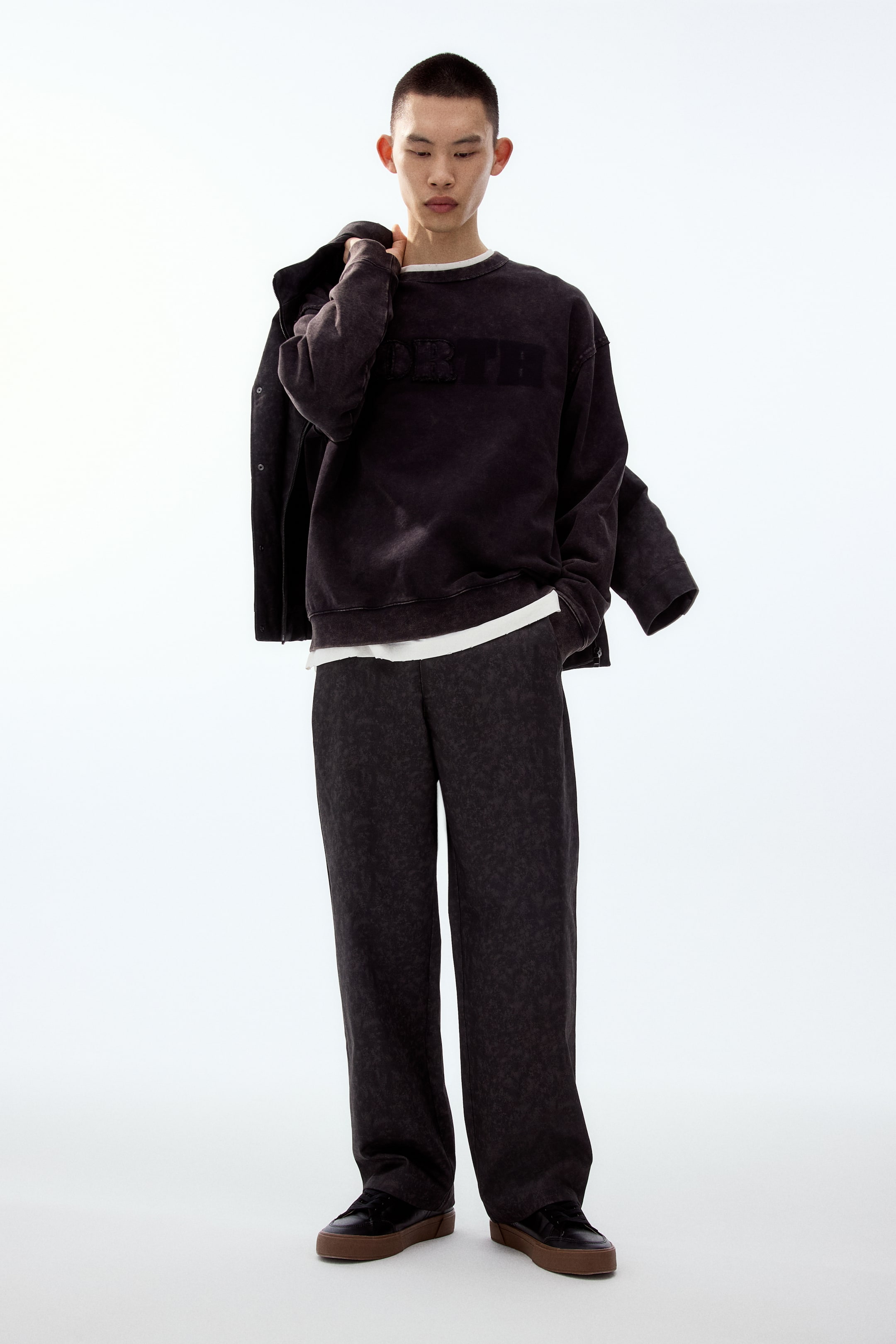 拡大画像を表示: ダークでウォッシュ加工の施されたクルーネックスウェットは、胸元に立体的で同系色のロゴを配し、袖口と裾はリブ仕様。白いTシャツをレイヤードしています。合わせたワイドパンツは、控えめなダークテクスチャの柄をあしらい、リラックスフィットのシルエットです。