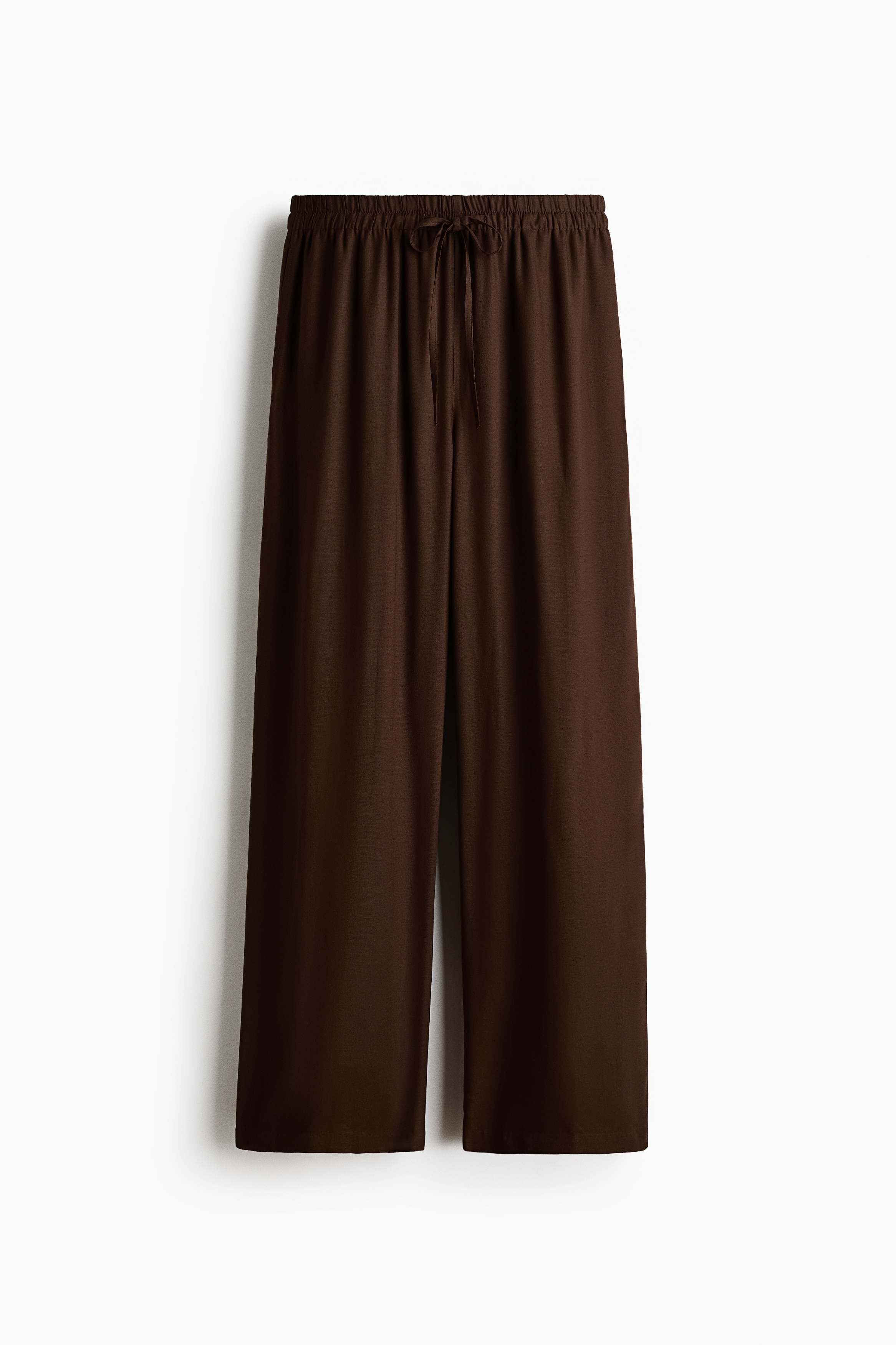 Brown loose-fit trousers featuring an elasticated drawstring waist and wide legs are being worn by the model. The trousers are paired with a matching open brown button-up shirt and a white top.