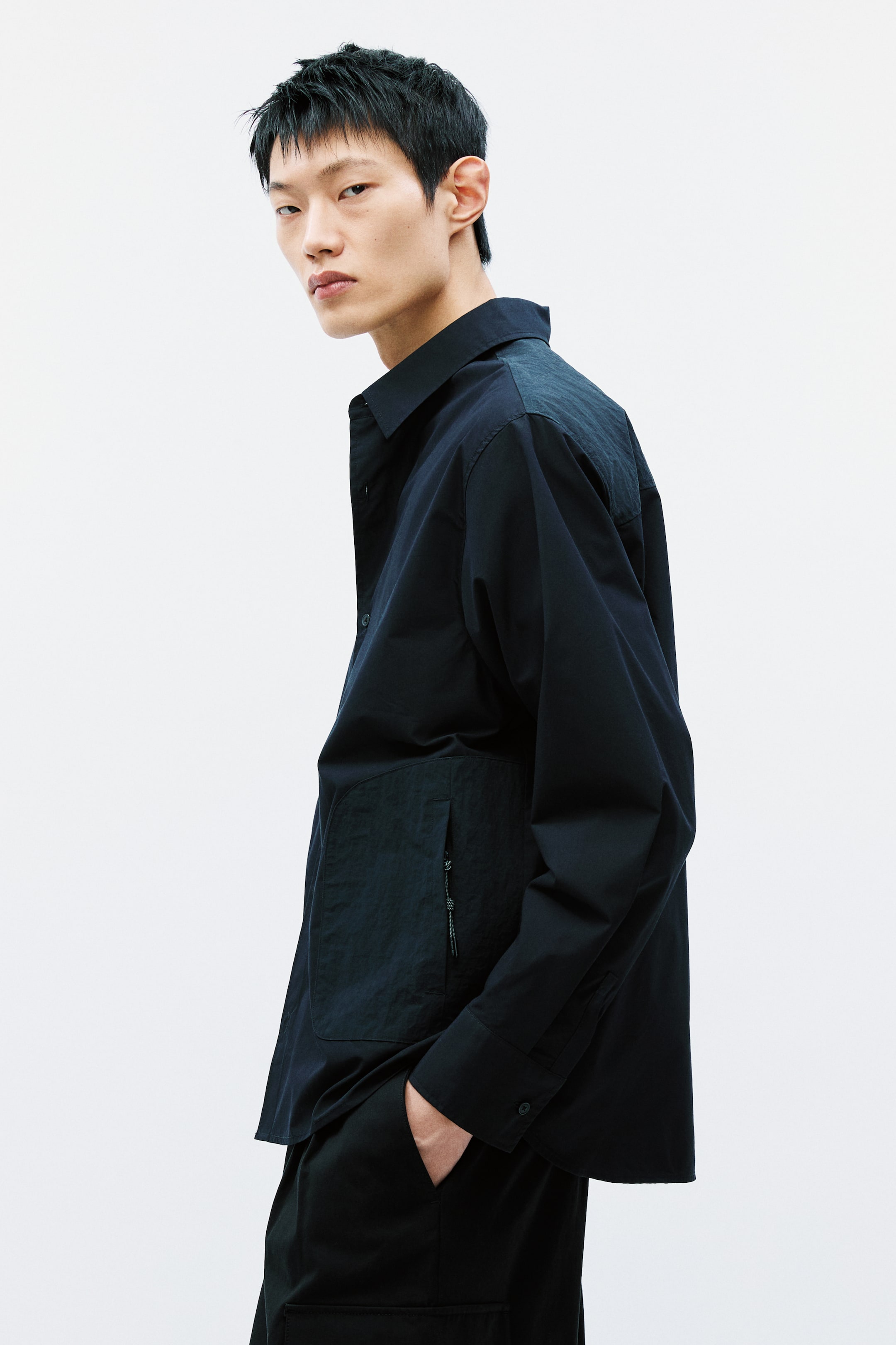 이미지 확대 보기: 검정색 긴소매 셔츠는 표준 칼라, 버튼 커프스, 버튼이 달린 앞면을 특징으로 합니다. 왼쪽 앞면에는 세로 지퍼가 있는 텍스처 패치 포켓이 있으며, 어깨를 가로지르는 동일한 텍스처 요크가 있습니다.