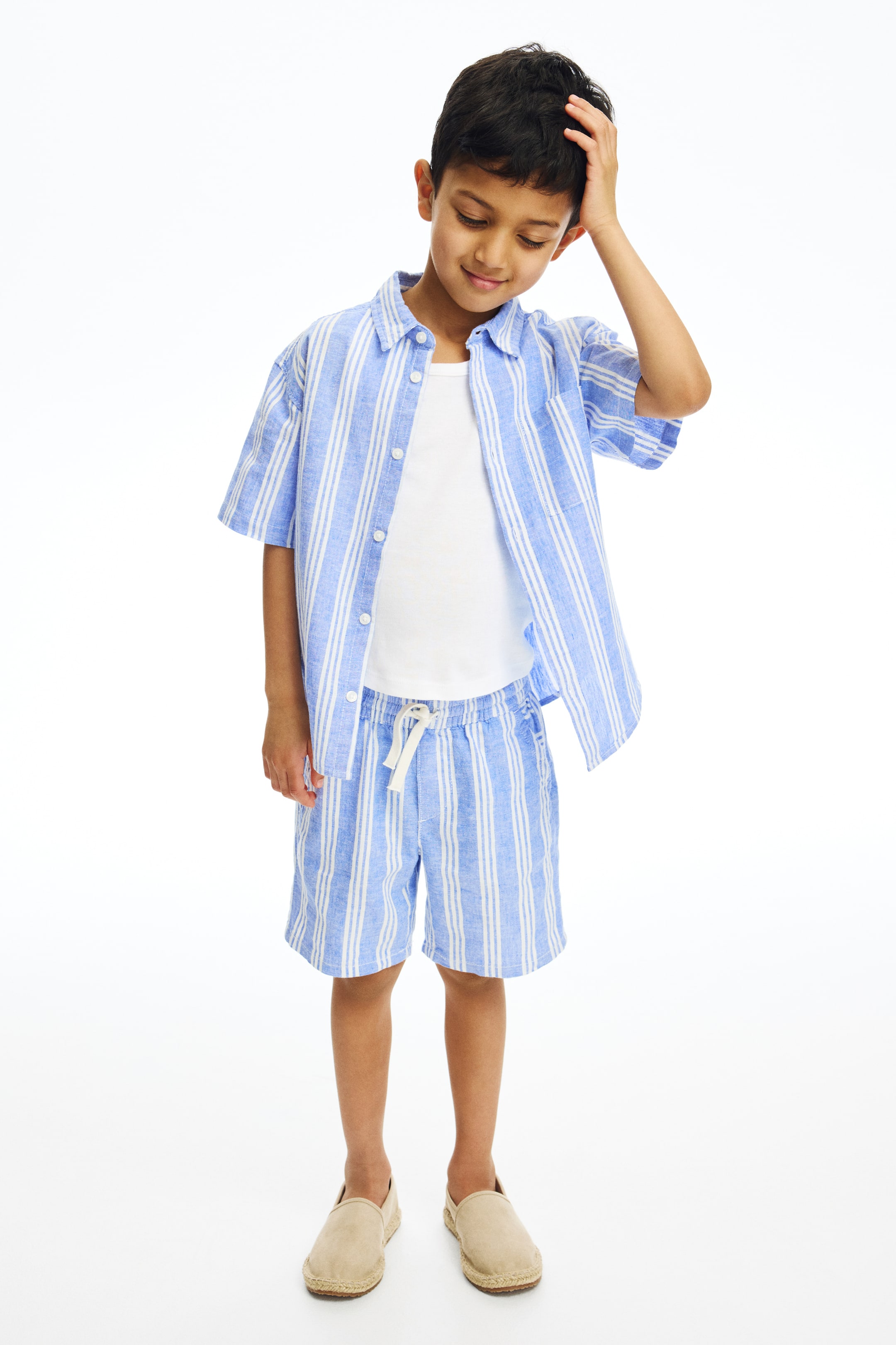 Vis større bilde: Et lyseblått og hvitt stripete kortermet skjorte- og shortssett presenteres, vendt forover. Skjorten har en krage og bæres åpen over en hvit t-skjorte, mens shortsen har en elastisk snor i livet.