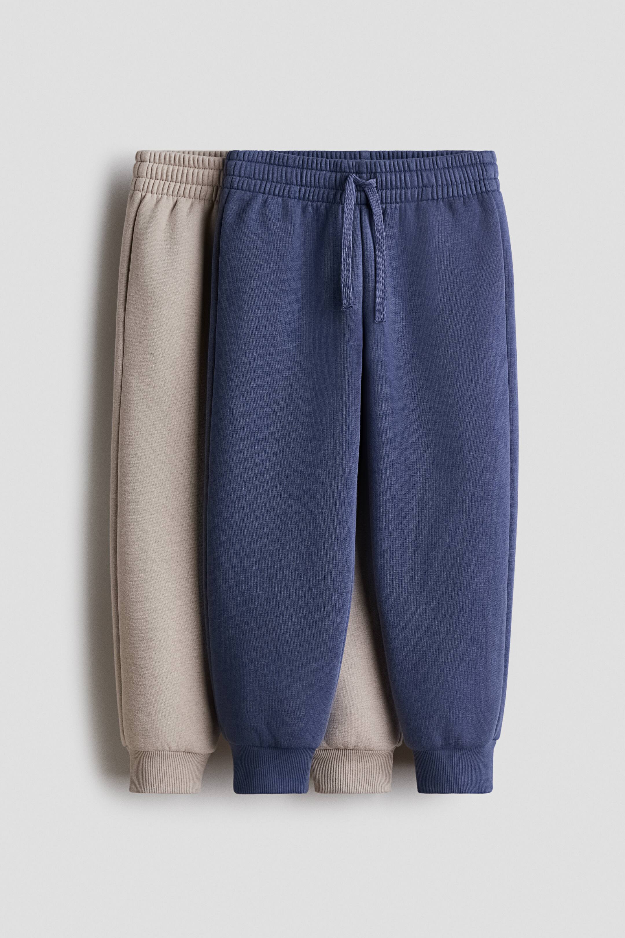 View larger image: Two pairs of joggers are facing FRONT, one dark blue and one beige, with the blue pair positioned slightly in front. Both are featuring elasticated, drawstring waists with a visible drawstring on the blue pair, side pockets, and ribbed trim at the hems, appearing in a sweatshirt fabric.