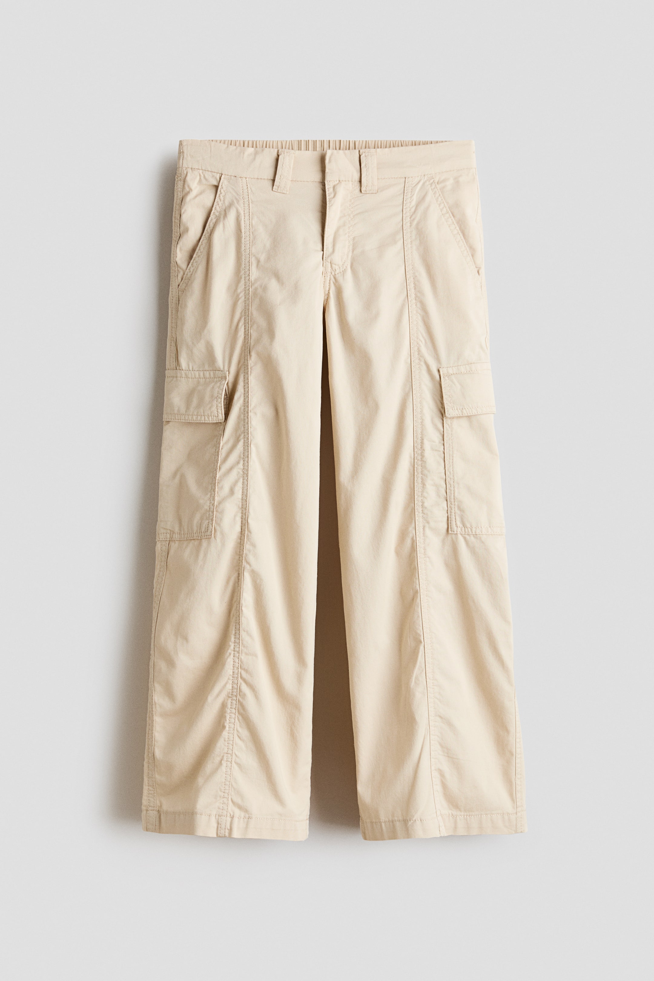 View larger image: Light beige cargo pants are showcased in a front view, featuring an elasticated back waistband, belt loops, slanted side pockets, and large flap cargo pockets on each leg. The wide-leg pants also display vertical stitched seams and subtle ruching down the front and lower legs.