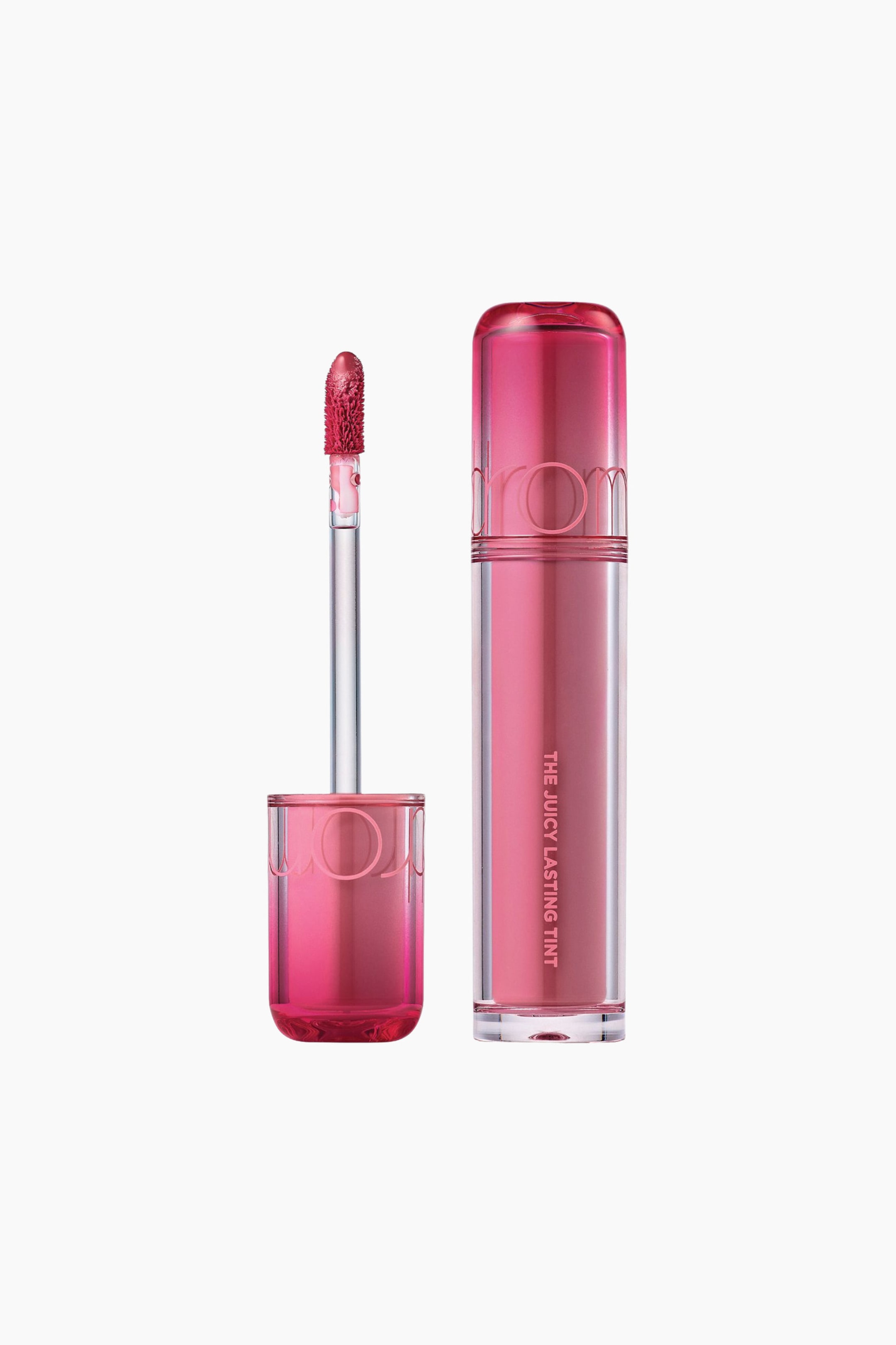 Vis større bilde: Et lip tint-produkt presenteres i to varianter: ett lukket og ett åpnet med applikatoren. Den lukkede, gjennomsiktige sylindriske flasken viser en rosa-bærfarget væske inni, med en halvtransparent rosa kork som har «ROMAND» og «THE JUICY LASTING TINT» trykt vertikalt. Det åpne produktet har den klare applikatorpinnen stående oppreist, med en floket tupp mettet med samme rosa-bærfarge, ved siden av den gjennomsiktige sylindriske basen.