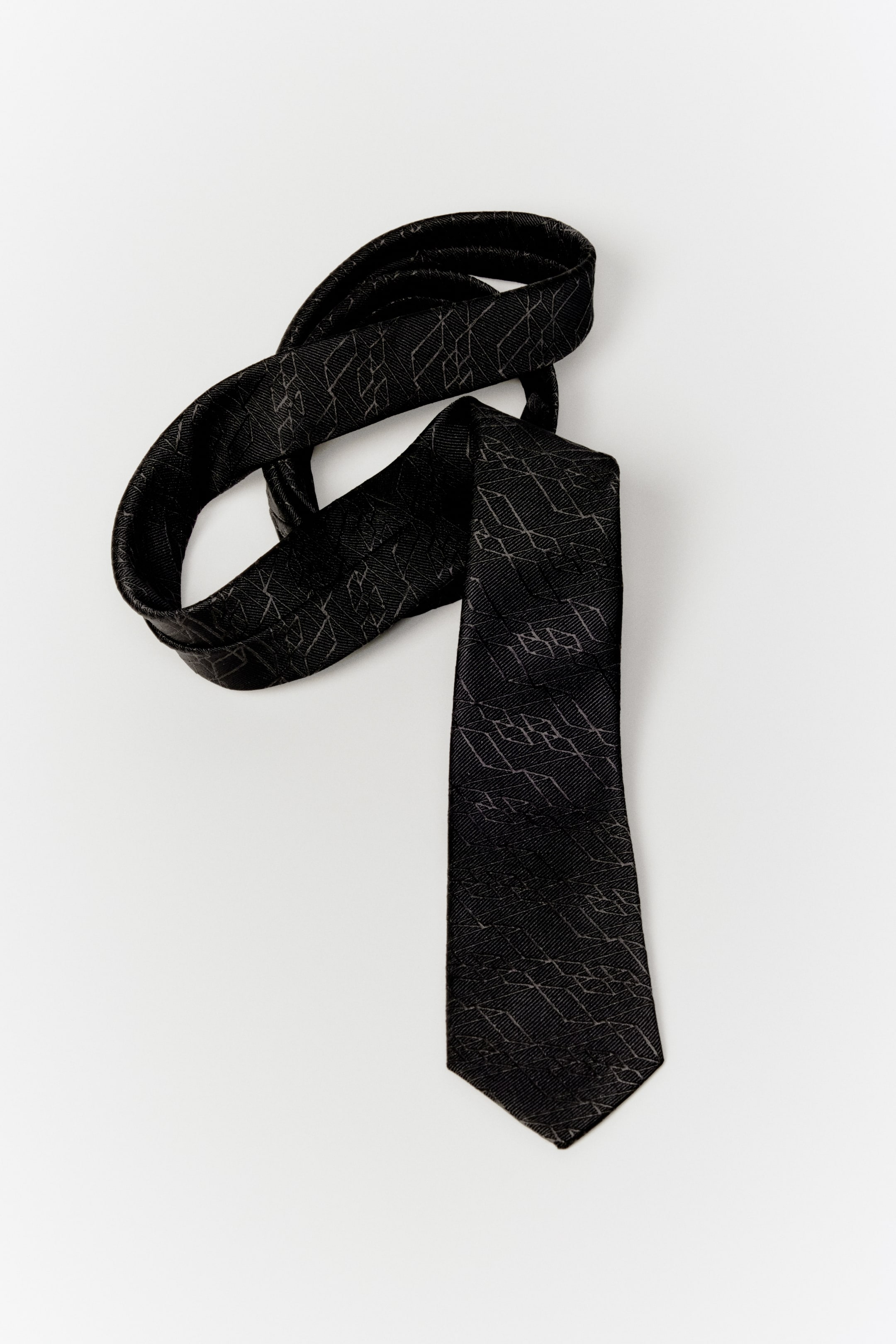View larger image: A black necktie is laid out with its wider end pointing downward, showcasing a subtle dark gray geometric pattern of interlocking angular shapes. The narrower end of the tie is looped upward.