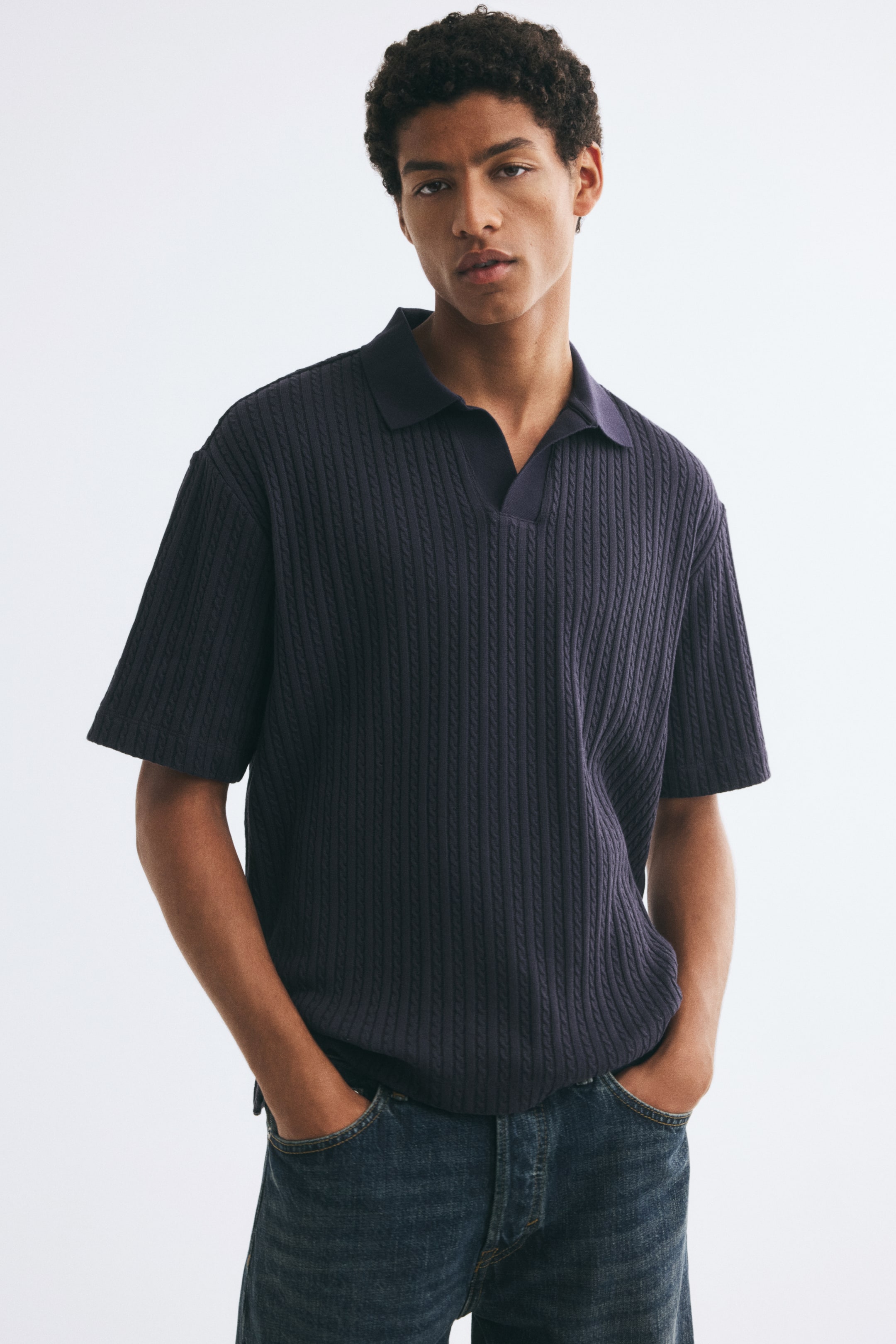 View larger image: A dark navy blue cable-knit polo shirt featuring an open collar, short sleeves, and a relaxed fit is being worn by the model. The shirt is styled with blue denim jeans, with the model standing with hands in their pockets.